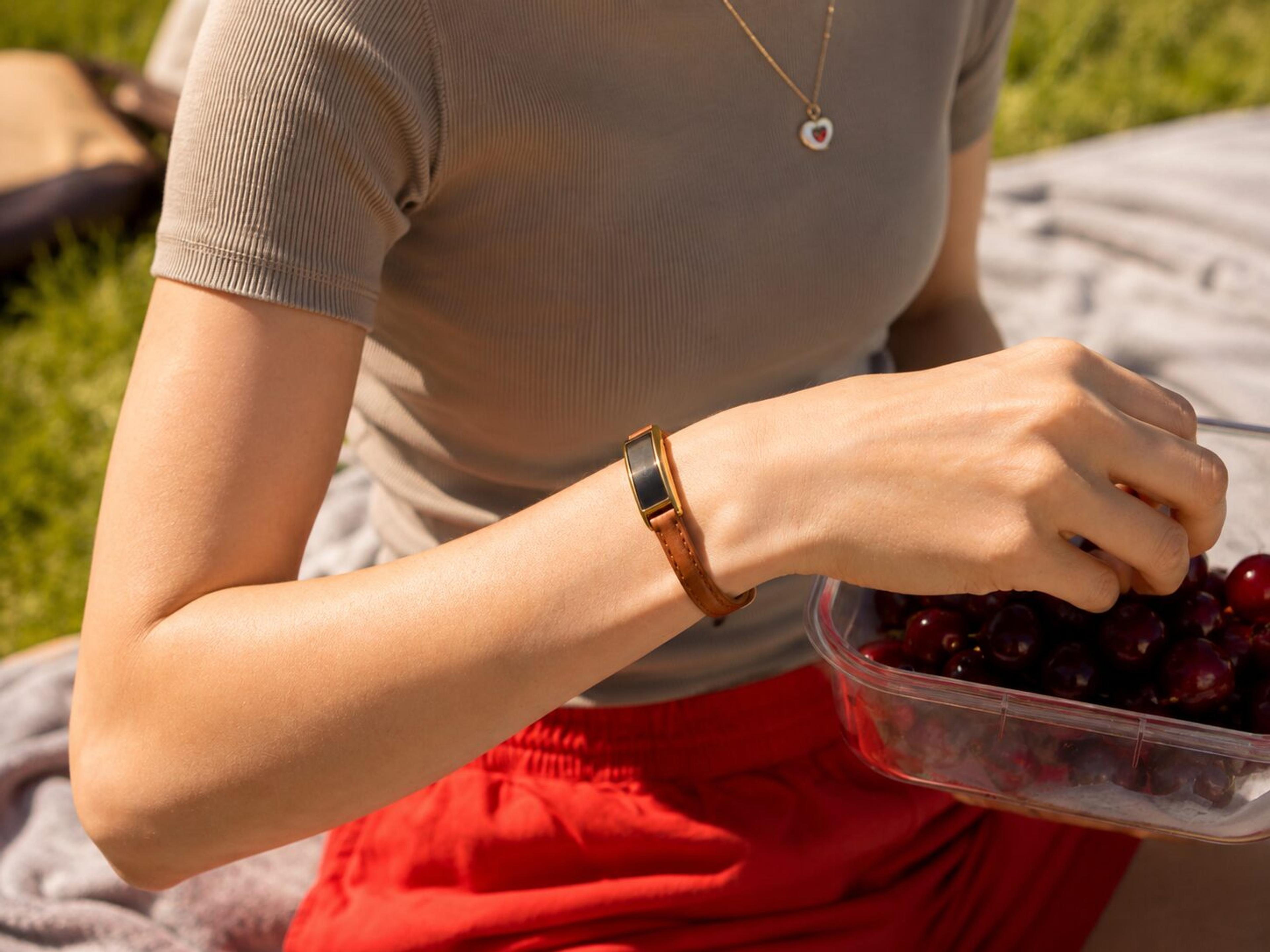 A person wearing a brown leather bracelet picks cherries from a container outdoors.