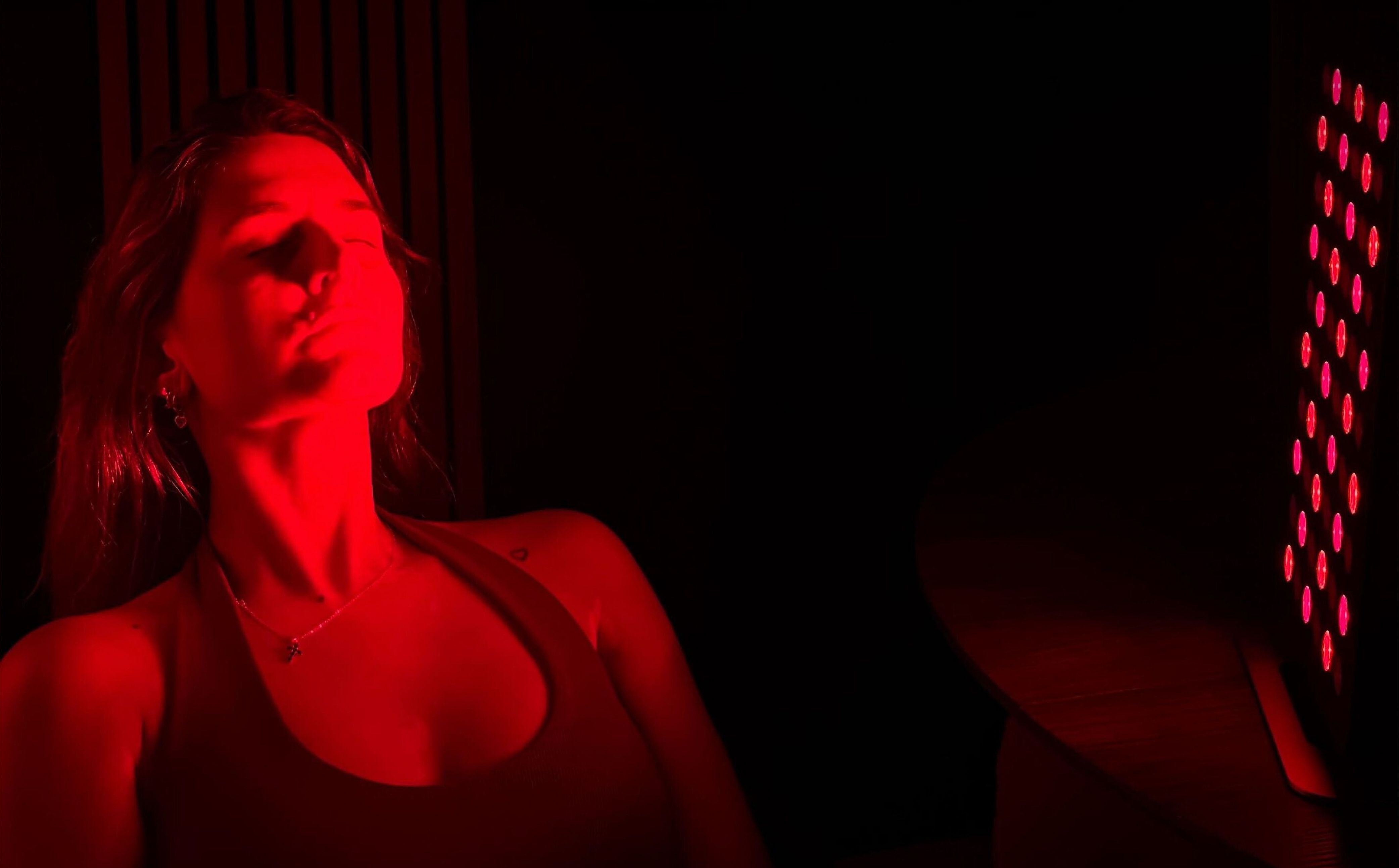 A woman with her eyes closed, illuminated by red light from a therapy panel.