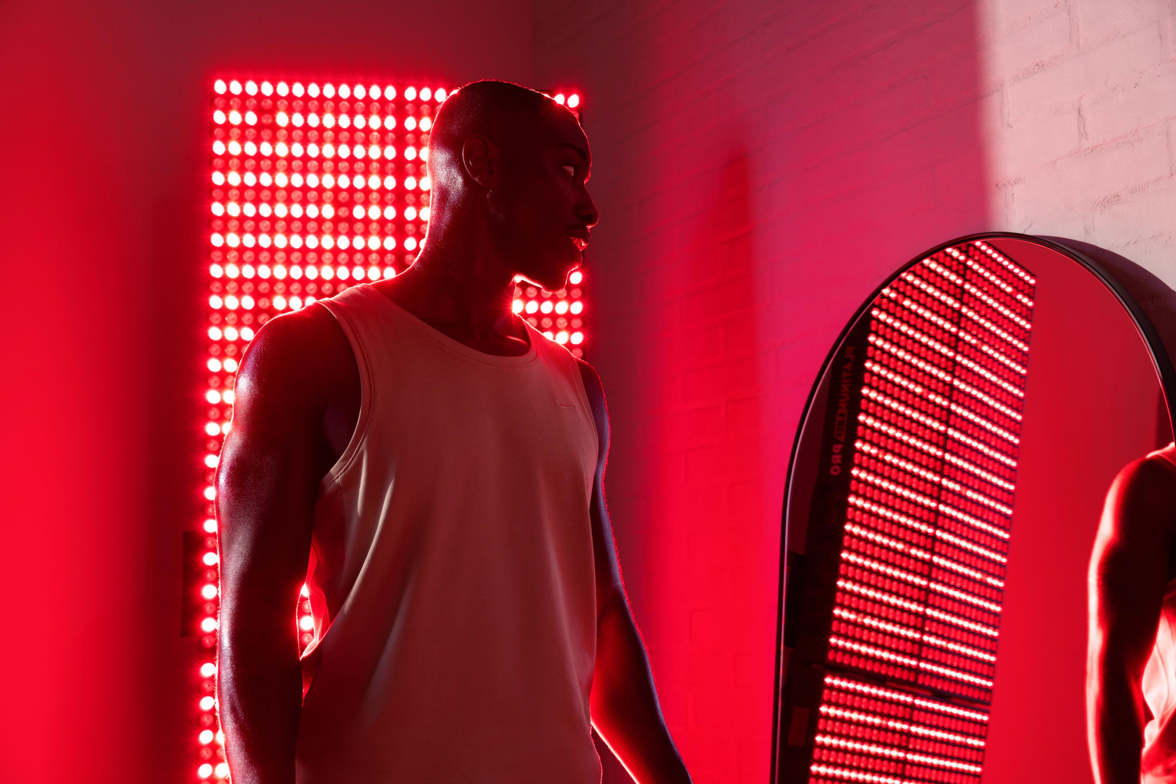 A man in a tank top stands beside a glowing vertical red light panel, which is reflected in an arched mirror.