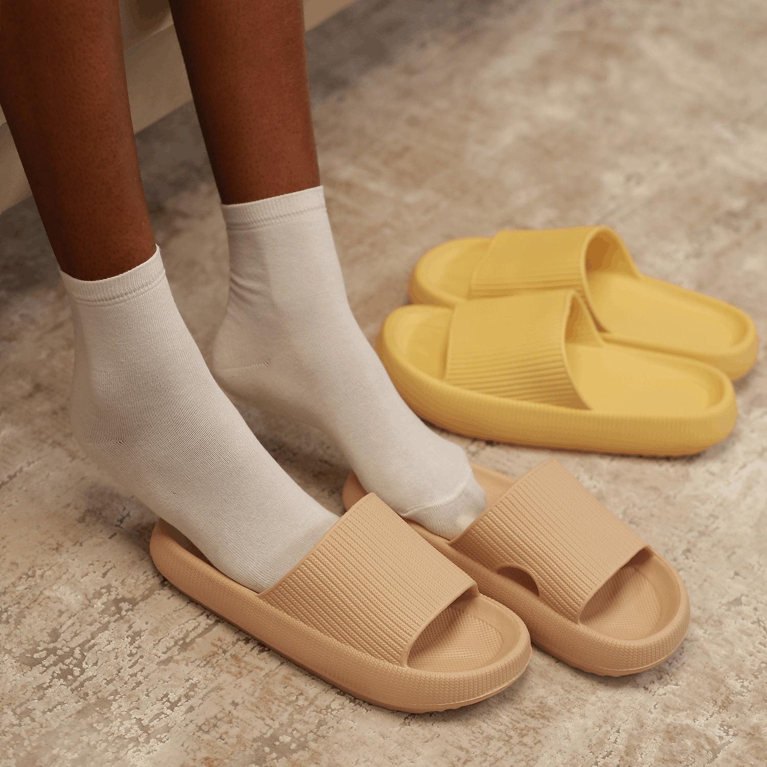 A person in white socks slides their foot into a beige sandal, with a pair of yellow slide sandals nearby on the floor.