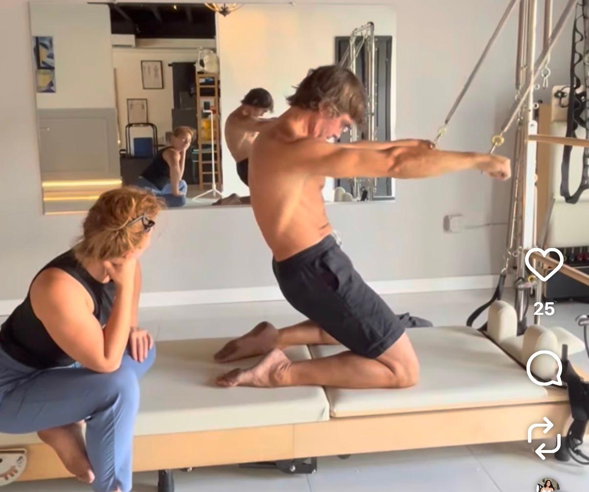 A shirtless man kneels on a Pilates reformer, holding straps and leaning forward, while a woman watches him intently.