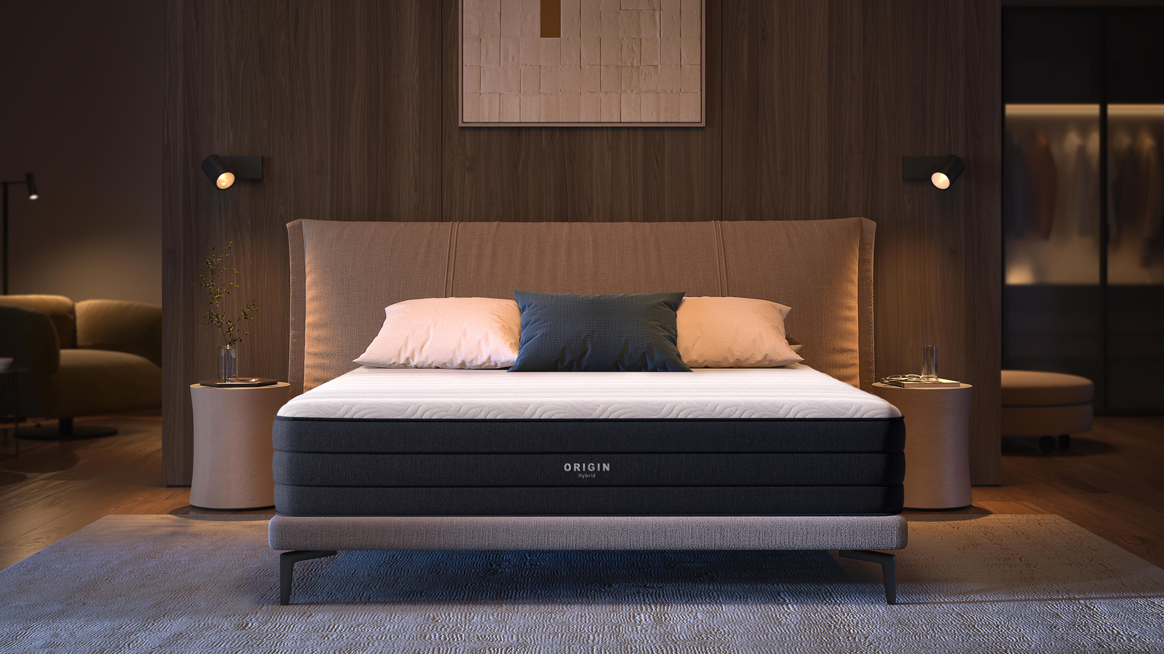 A black and white Origin mattress on a bed in a modern, warmly lit bedroom with a beige headboard and bedside tables.