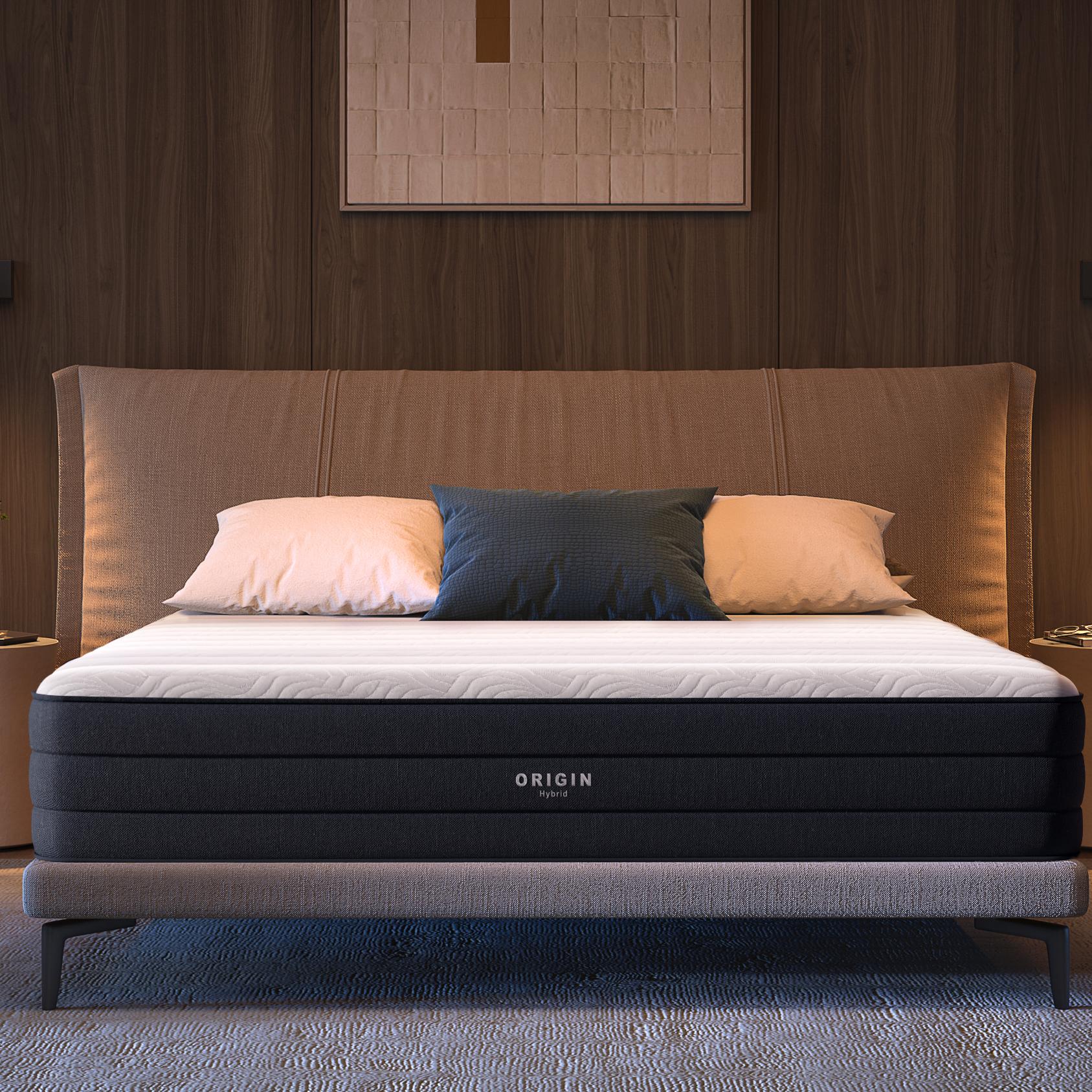 A black and white Origin mattress on a bed in a modern, warmly lit bedroom with a beige headboard and bedside tables.
