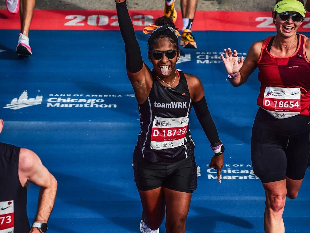 An excited runner raises her arm and sticks out her tongue at the Chicago Marathon finish line, with another smiling runner beside her.