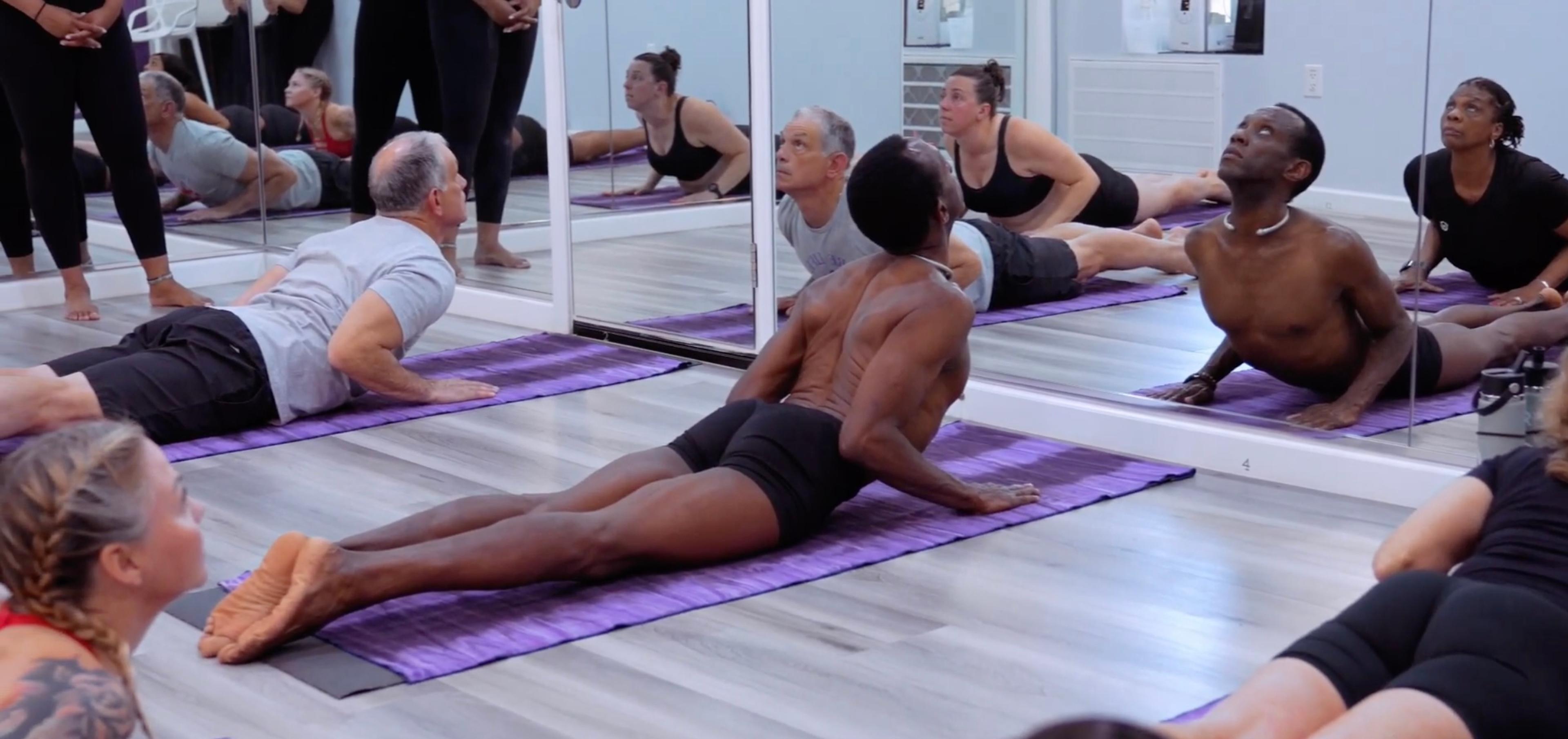 A diverse group of people in a fitness studio stand on purple mats, arms extended horizontally, facing a mirrored wall.