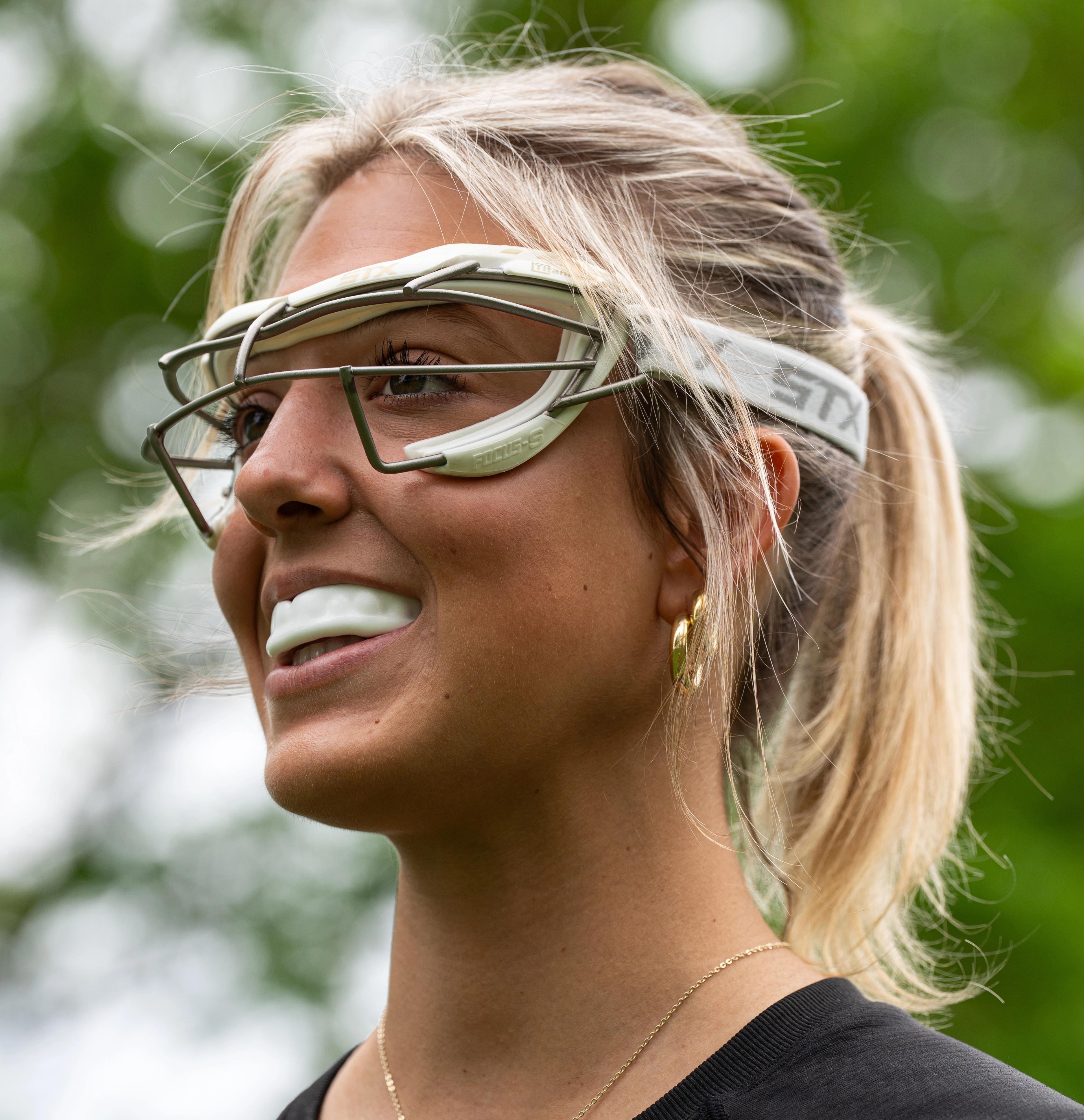A smiling young woman with a blonde ponytail wears lacrosse goggles and a mouthguard.