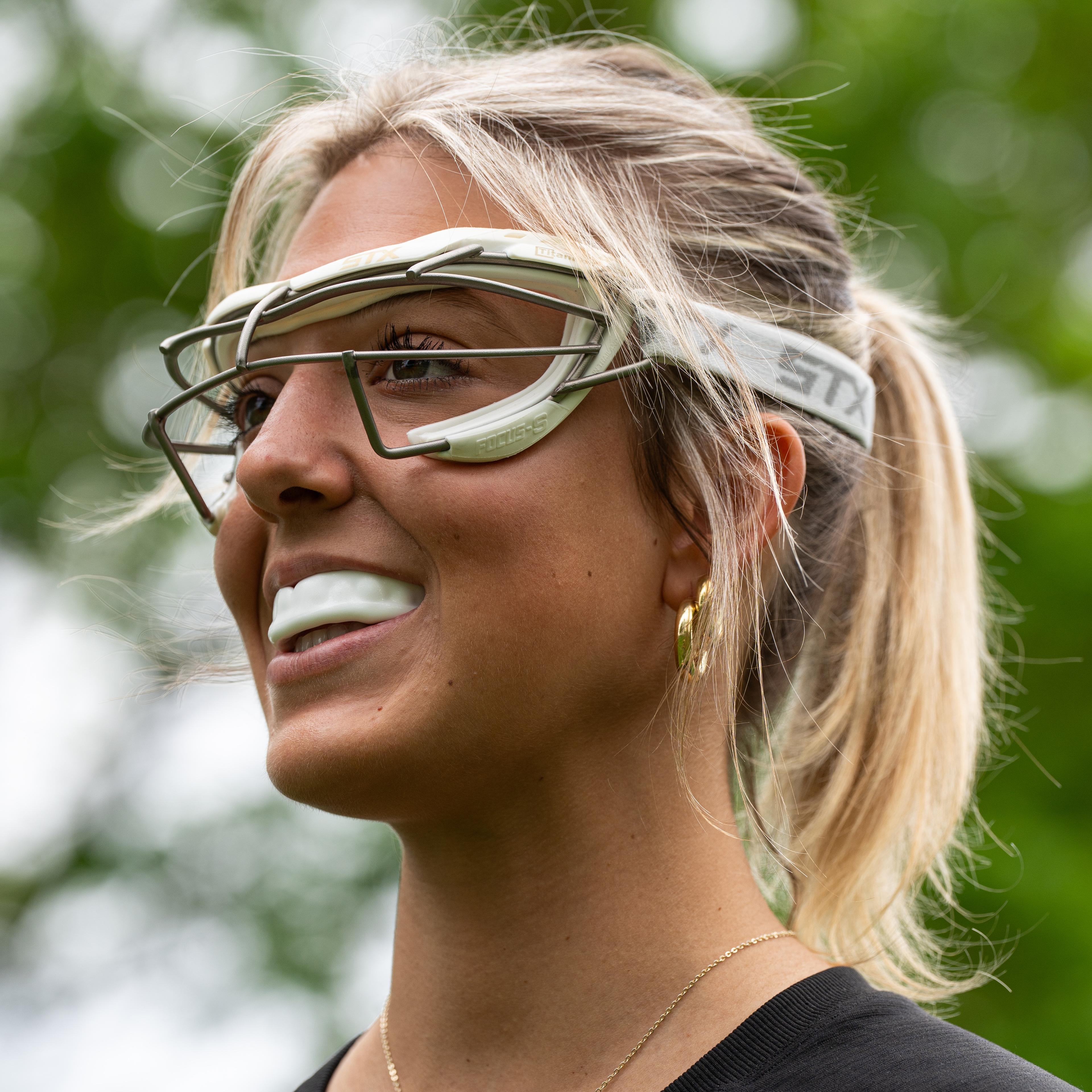 A smiling young woman with a blonde ponytail wears lacrosse goggles and a mouthguard.