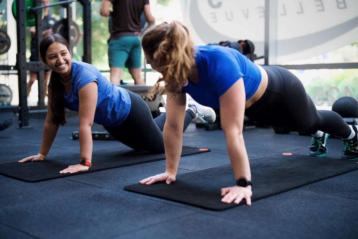 Two women in a gym doing planks on exercise mats, one smiling.
