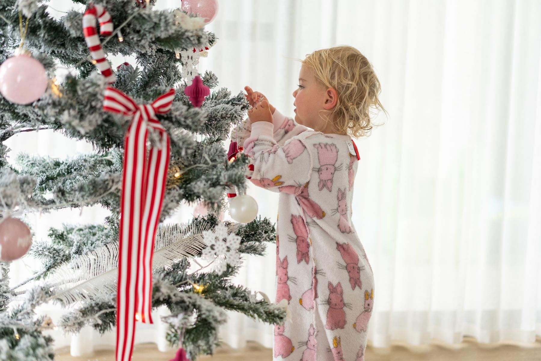 A toddler hanging decorations on a Christmas tree in front of white sheer curtains