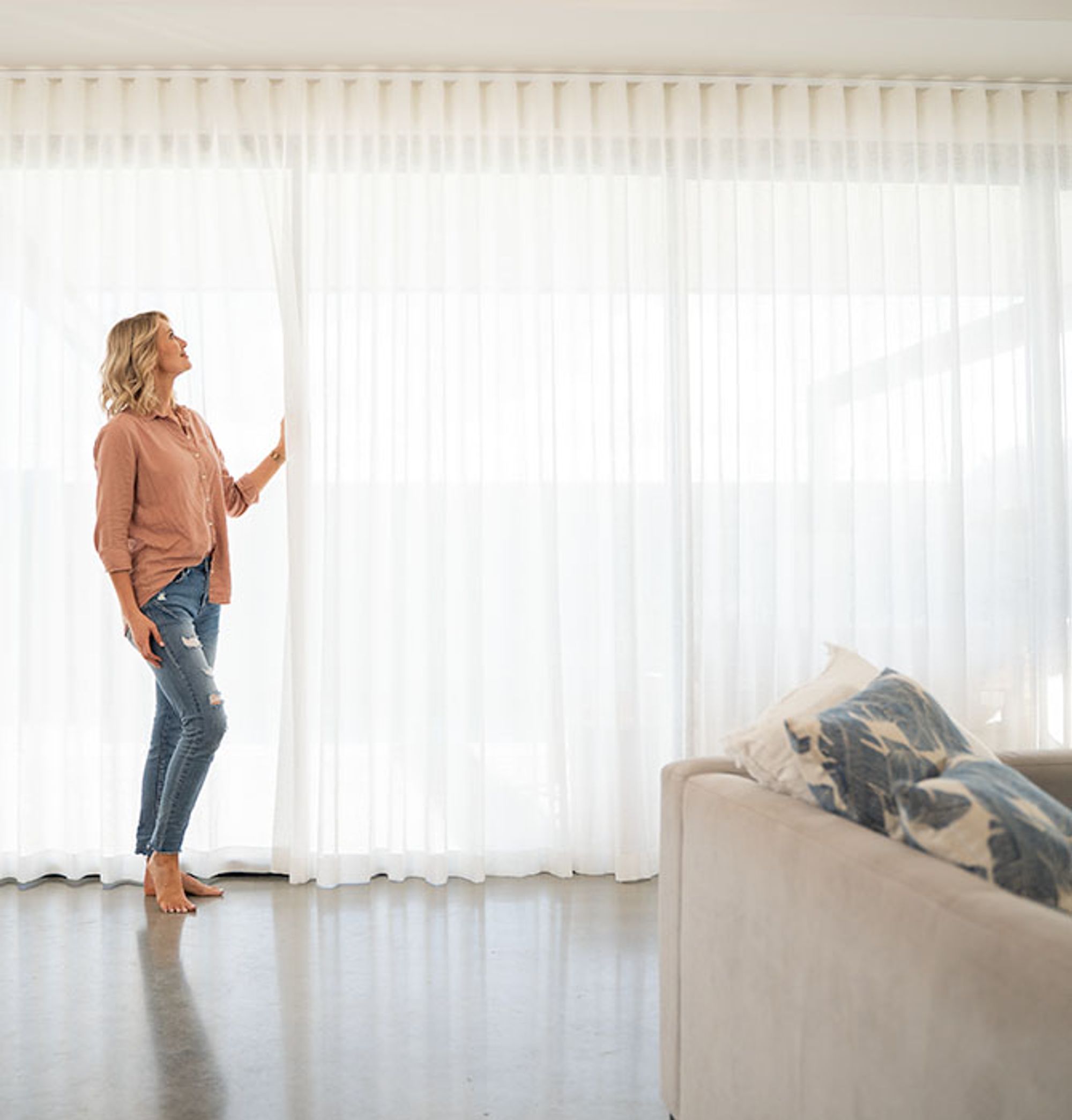A woman drawing floor-to-ceiling white sheer curtains in a living room