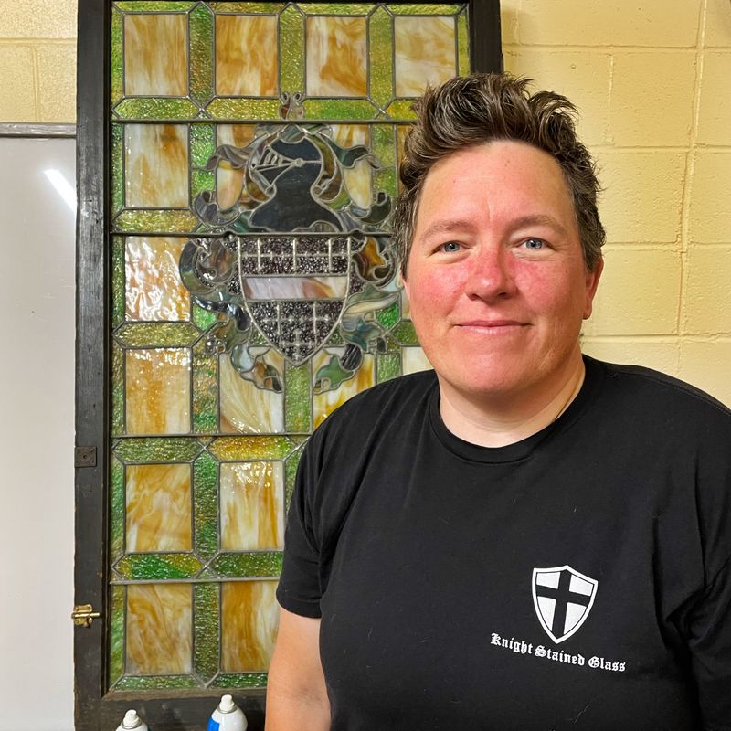 Owner Andrea Knight standing in front of a custom stained glass window at the Knight Stained Glass studio