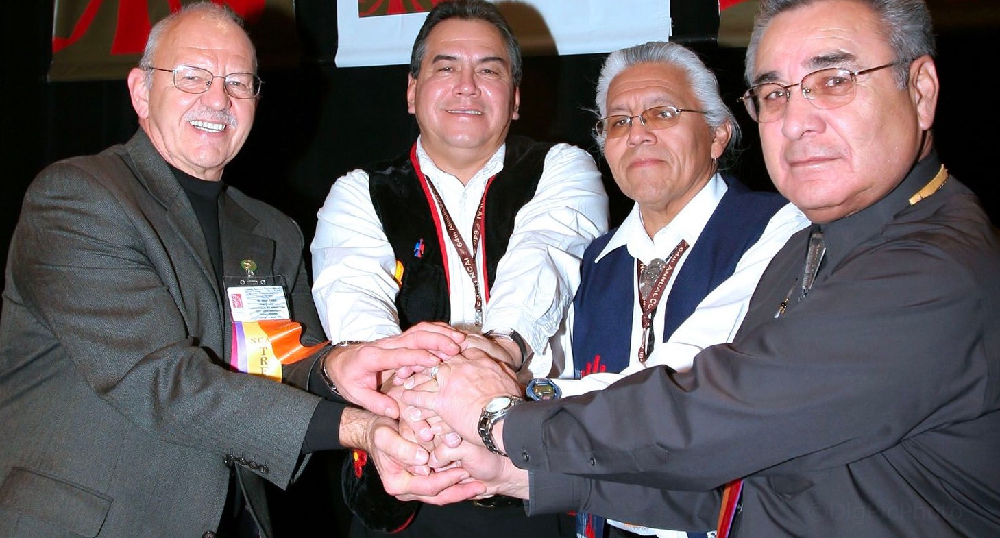 4 men put their hands together in front of a sign that reads "National Congress of American Indians"