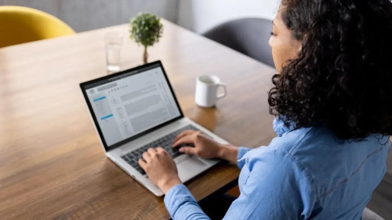 Woman working at a wooden desk on a laptop with an email inbox on screen, coffee mug and plants nearby