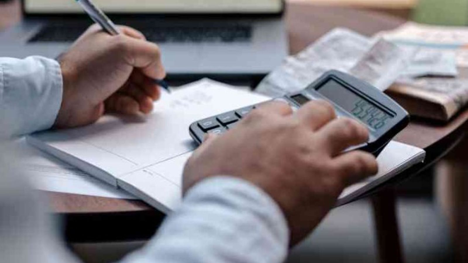Person using a calculator and writing in a notebook while working on invoices at a desk