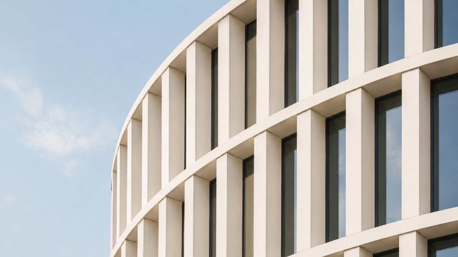 Modern curved building facade with repeating vertical columns and dark windows against a pale blue sky
