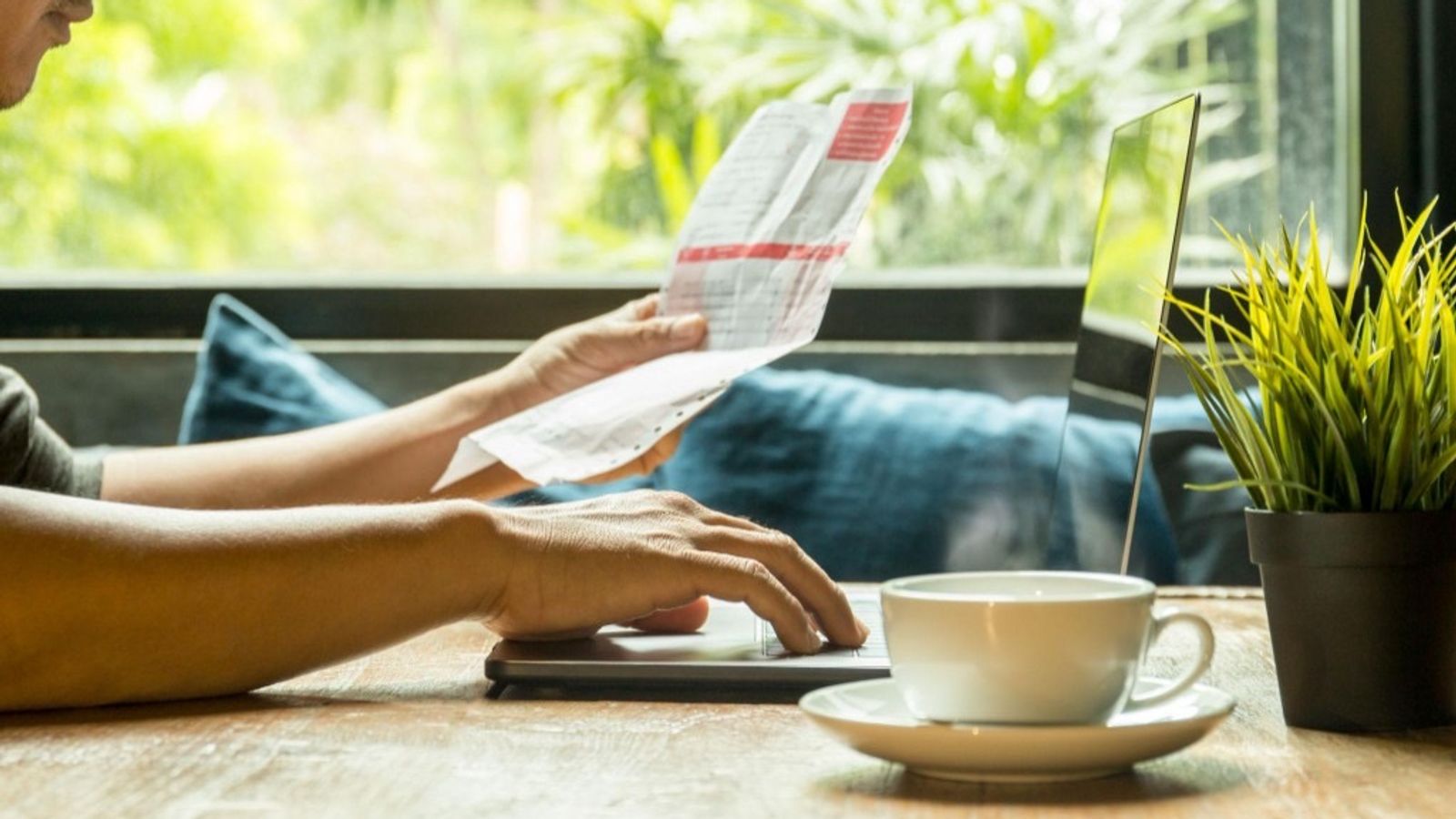 Person at a wooden desk reviewing a paper invoice while using a laptop, with coffee and a plant nearby