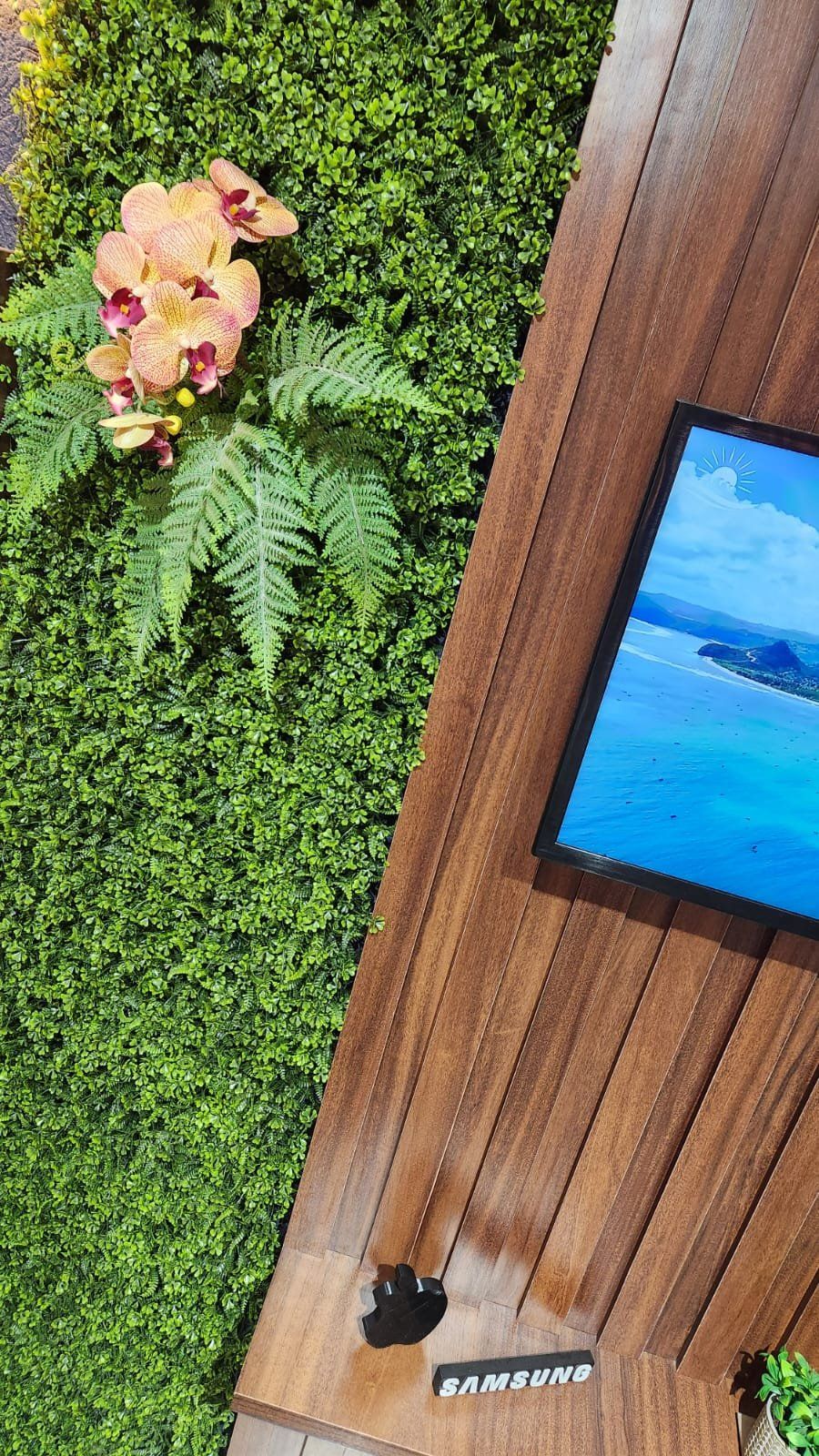 Detalhe do painel verde com orquídea ao lado da TV e do ripado de madeira