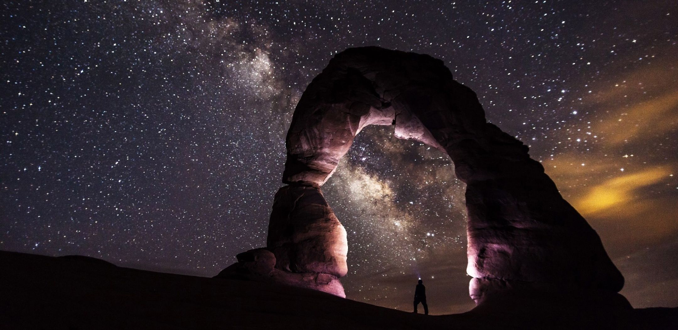 A starry night sky over a desert rock arch with a lone figure