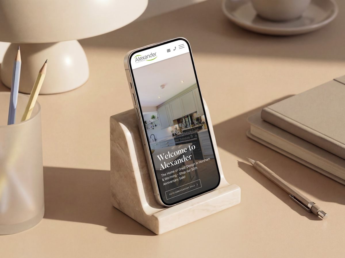 The Alexander Kitchens website on a phone, styled with a lamp and notebook on a warm surface
