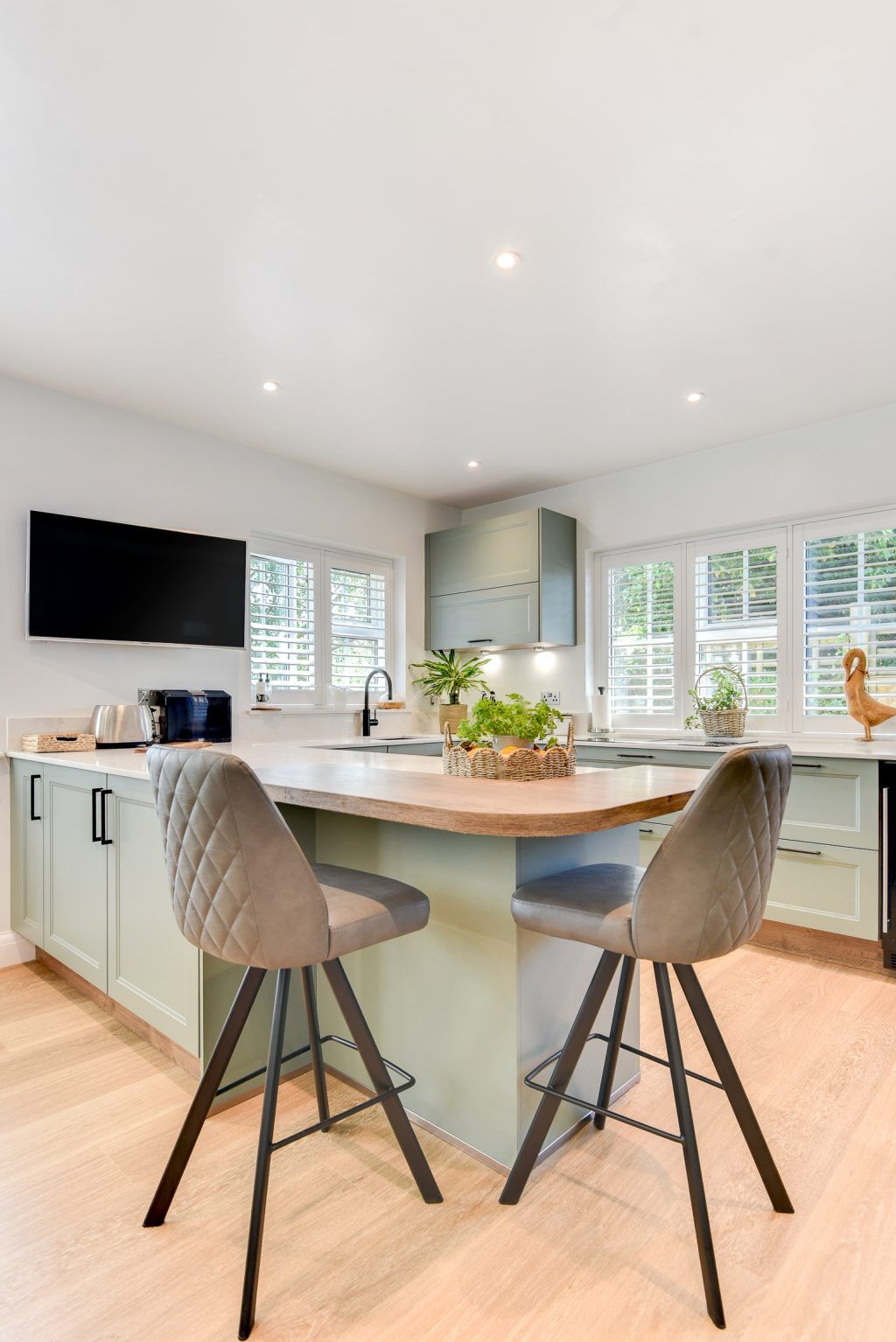 A bright sage-green kitchen with window shutters, a wall TV and bar stools