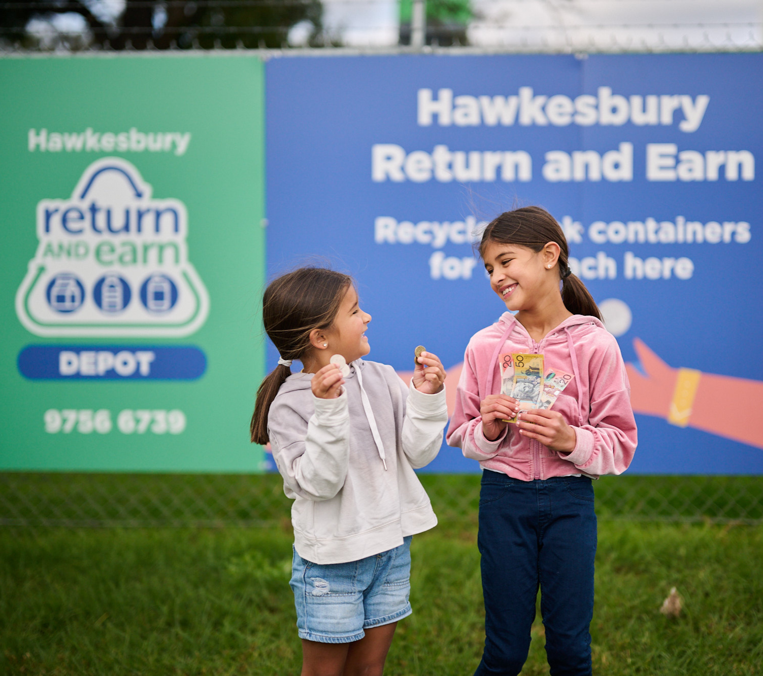 Two young girls are smiling and standing in front of a "Return and Earn" recycling depot sign. The girl on the left, wearing a light grey hoodie, is holding coins in her cupped hands. The girl on the right, dressed in a pink hoodie, is proudly holding up several Australian banknotes, including $5, $10, $20, and $50 notes. The background includes green and blue signage promoting container recycling in the Hawkesbury region.