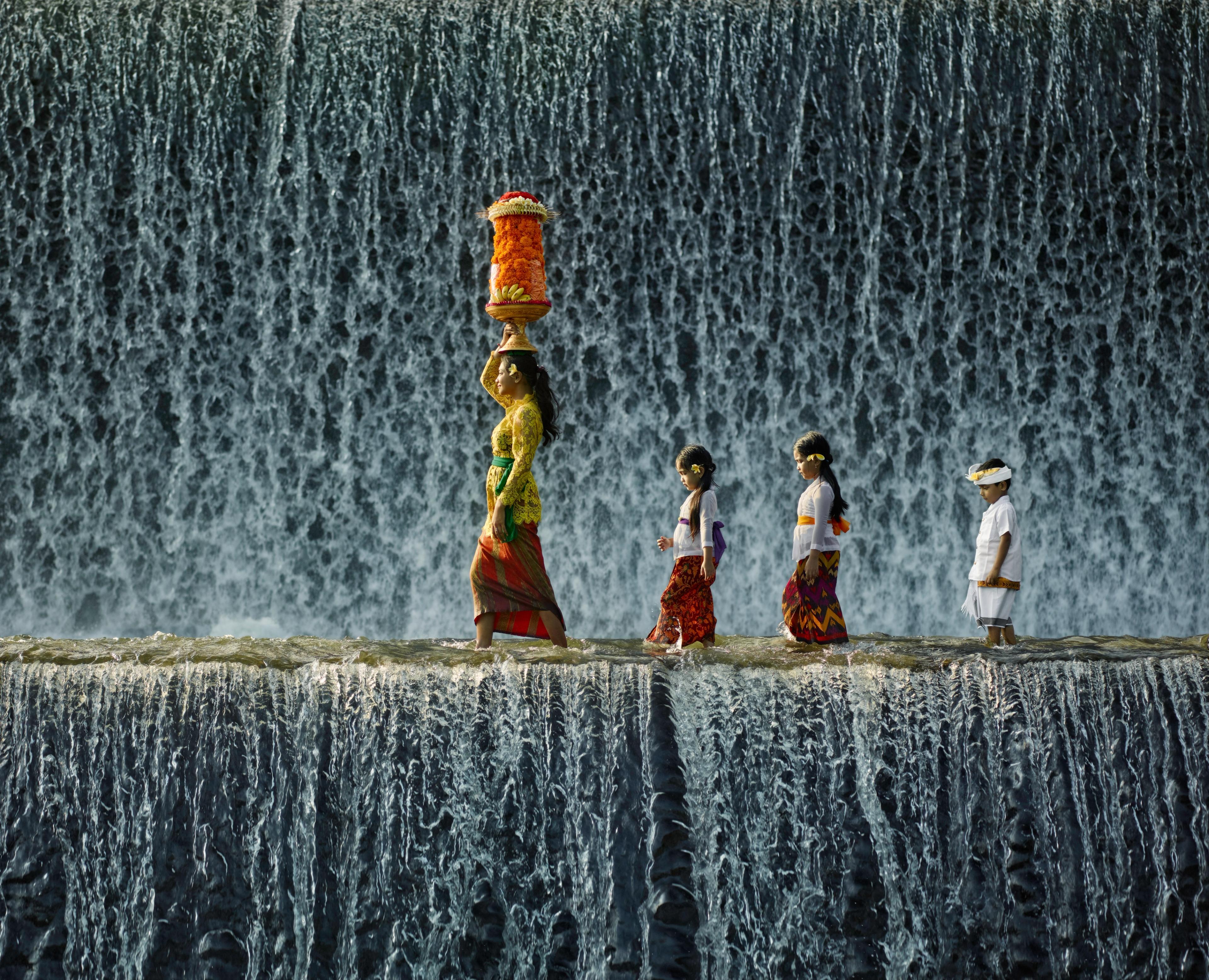 In Bali, a woman carries temple offerings, and children tag along across a waterfall in Bali/Getty images