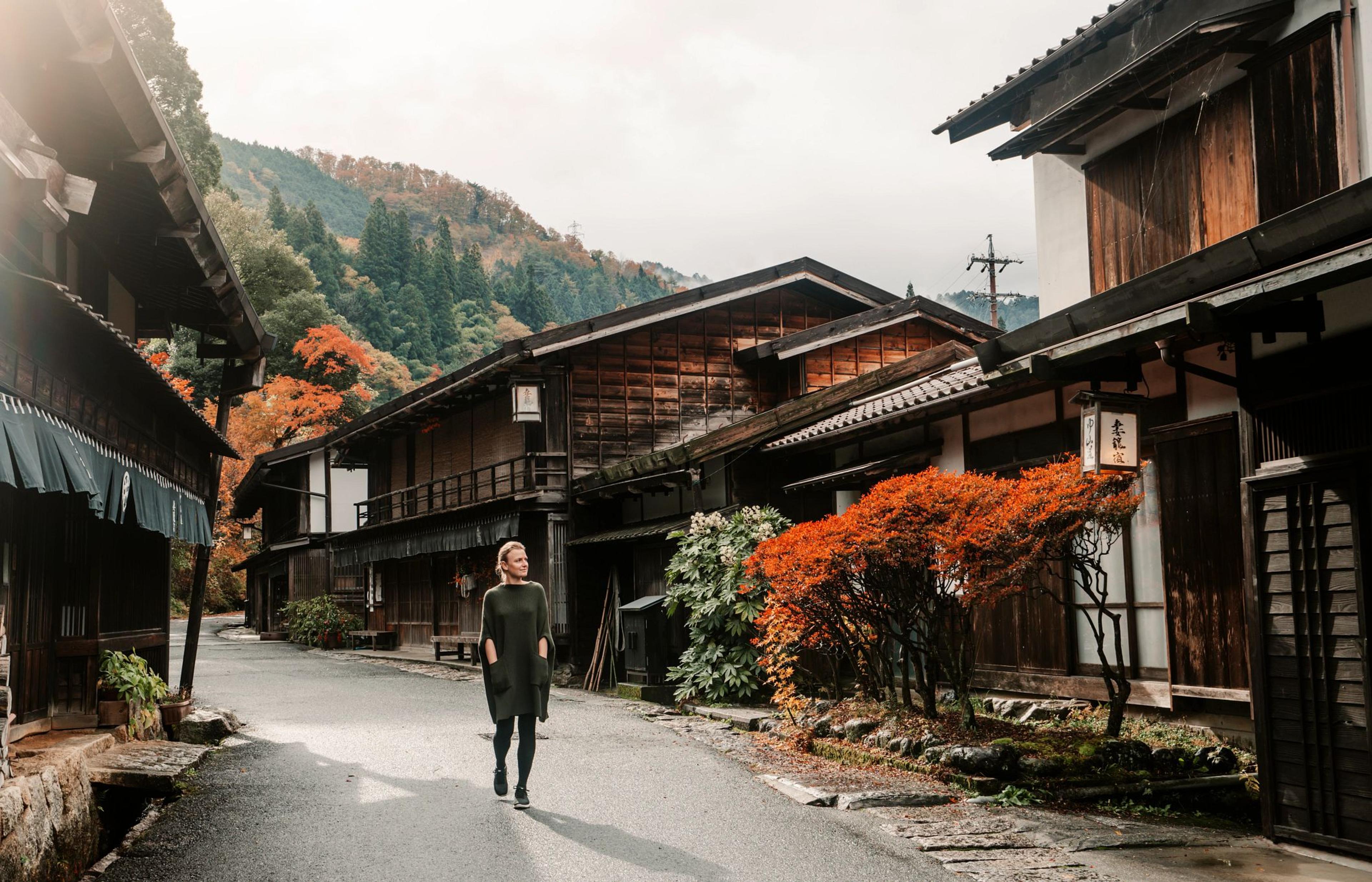 A traveler walks down a quiet street lined with traditional wooden buildings and vibrant autumn foliage in Tsumago-juku.