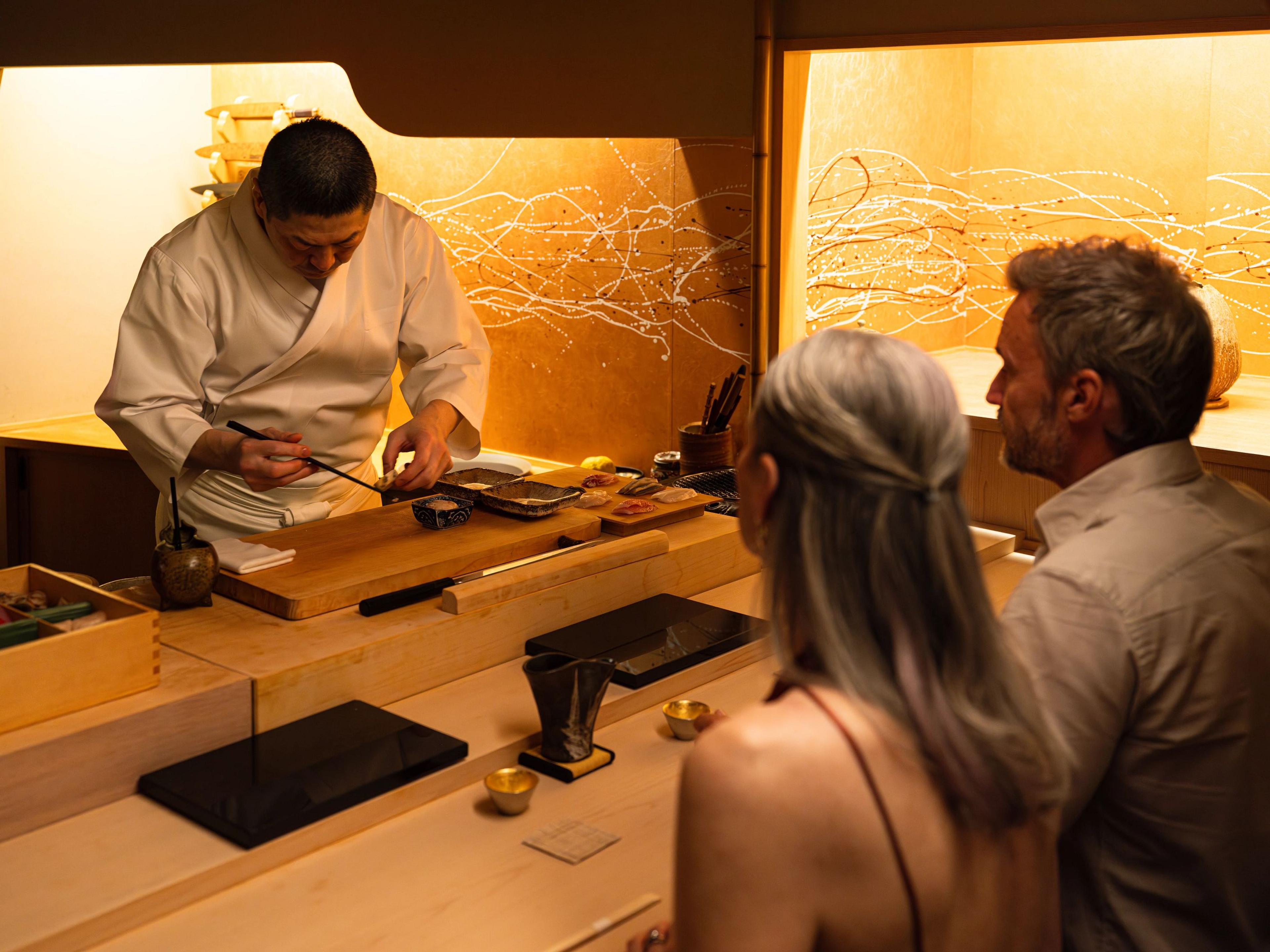 A chef meticulously prepares an omakase meal at a wooden counter for two seated guests. 
