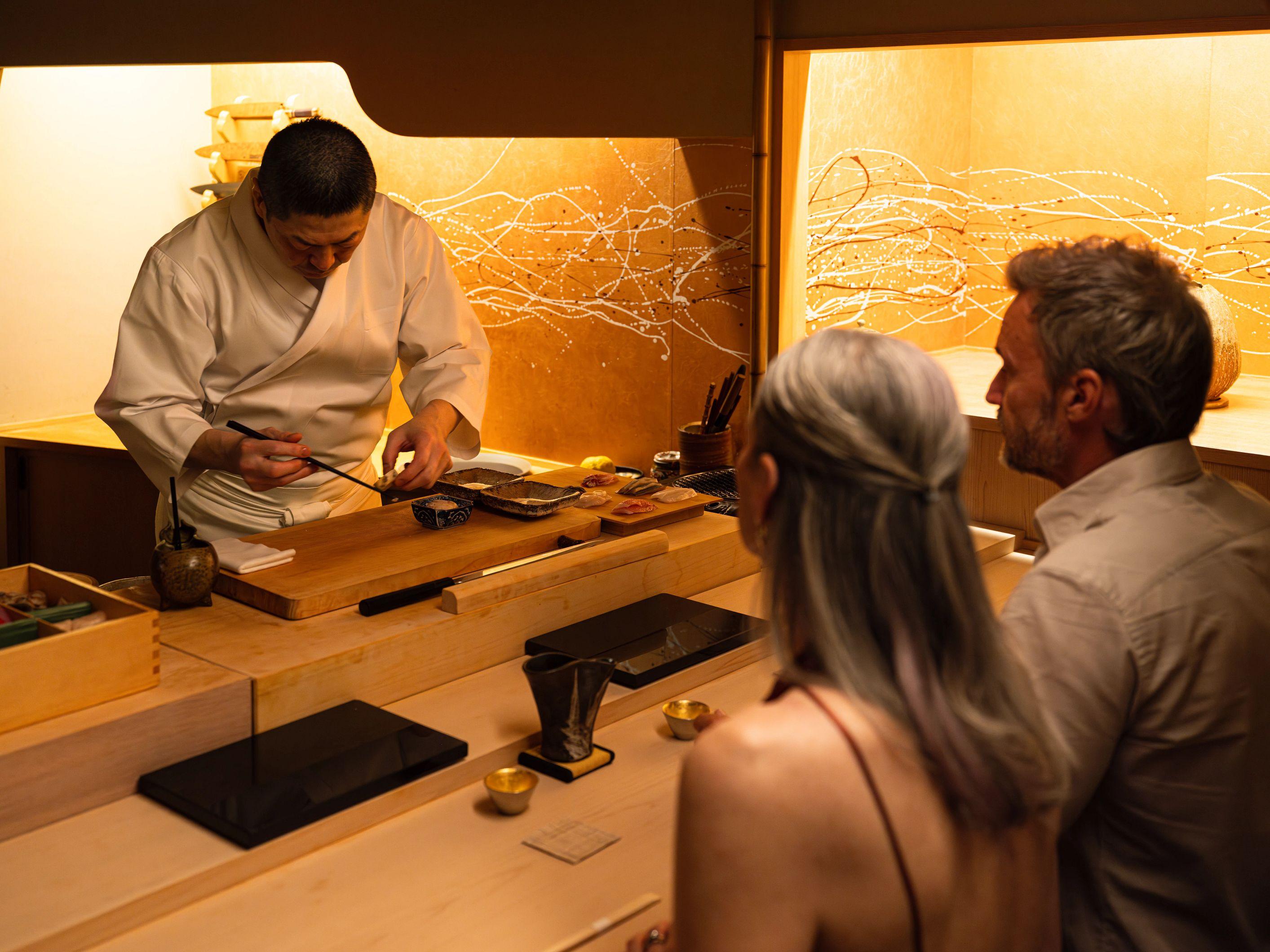 A chef meticulously prepares an omakase meal at a wooden counter for two seated guests.