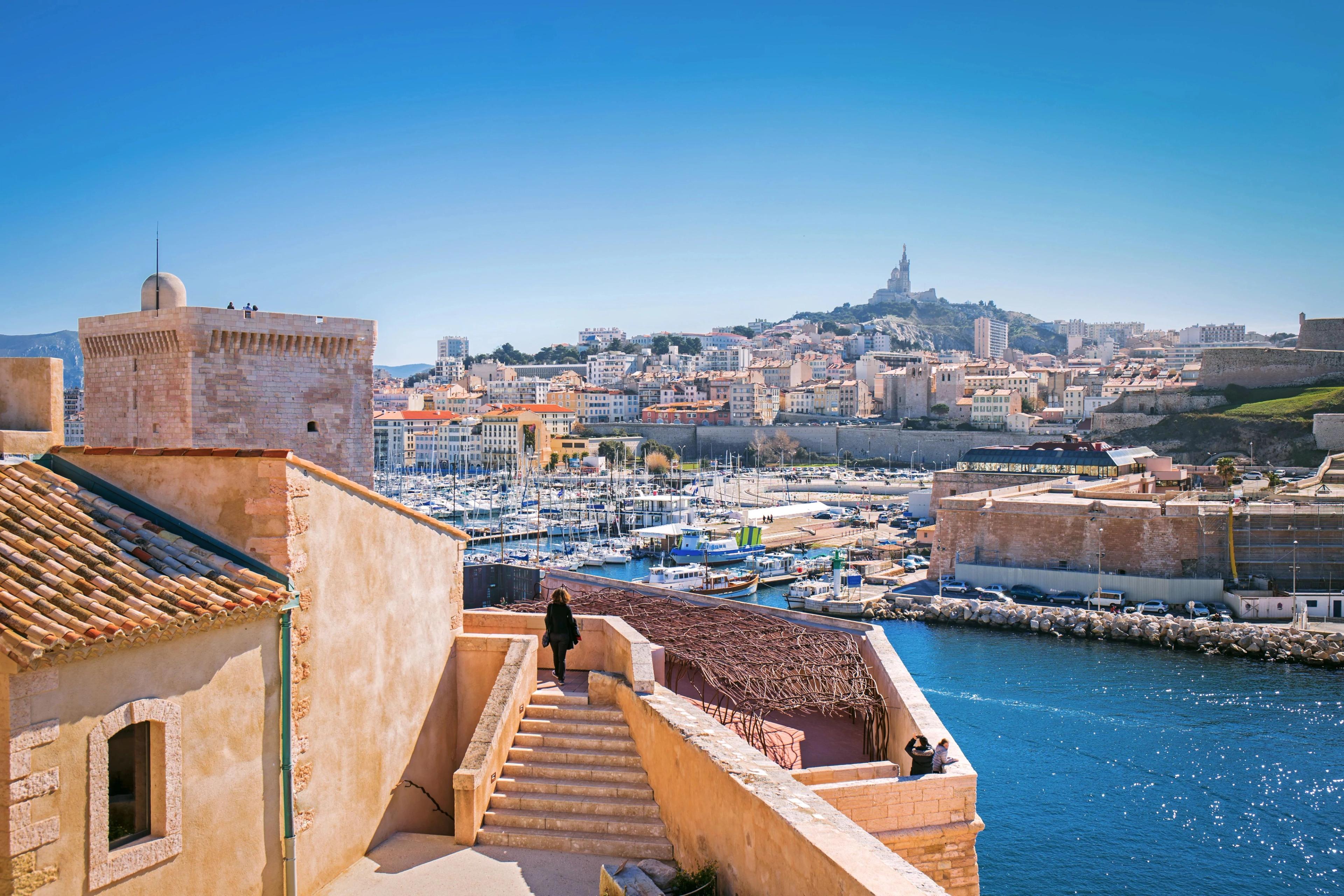 Marseille historic fort ramparts frame boat-filled Vieux-Port waters highlighting distant hillside basilica. 