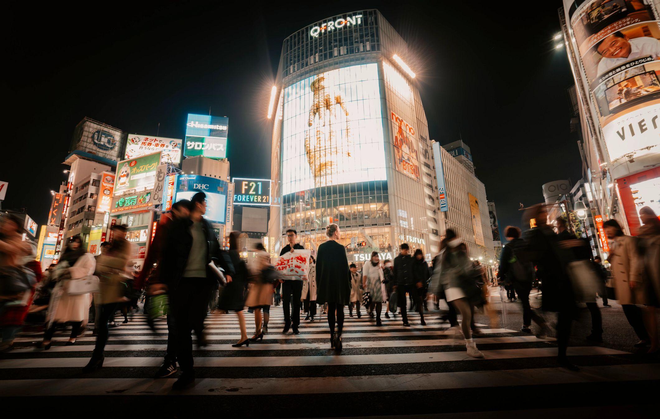 Crowds of pedestrians cross Tokyo's iconic Shibuya Crossing illuminated by glowing neon billboards at night.