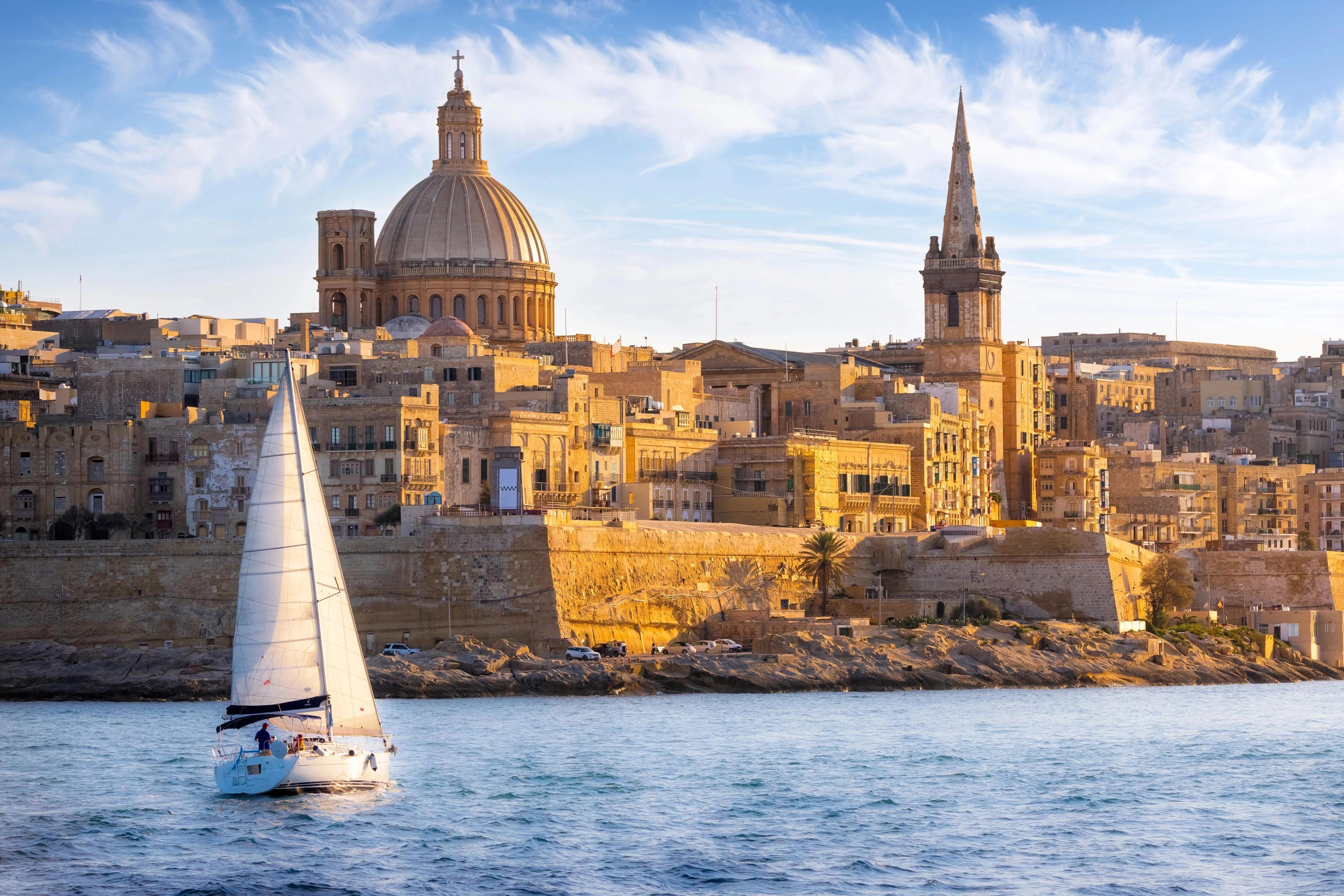 White sailboat cruising blue waters framing the historic Valletta Basilica dome and cathedral spire. 
