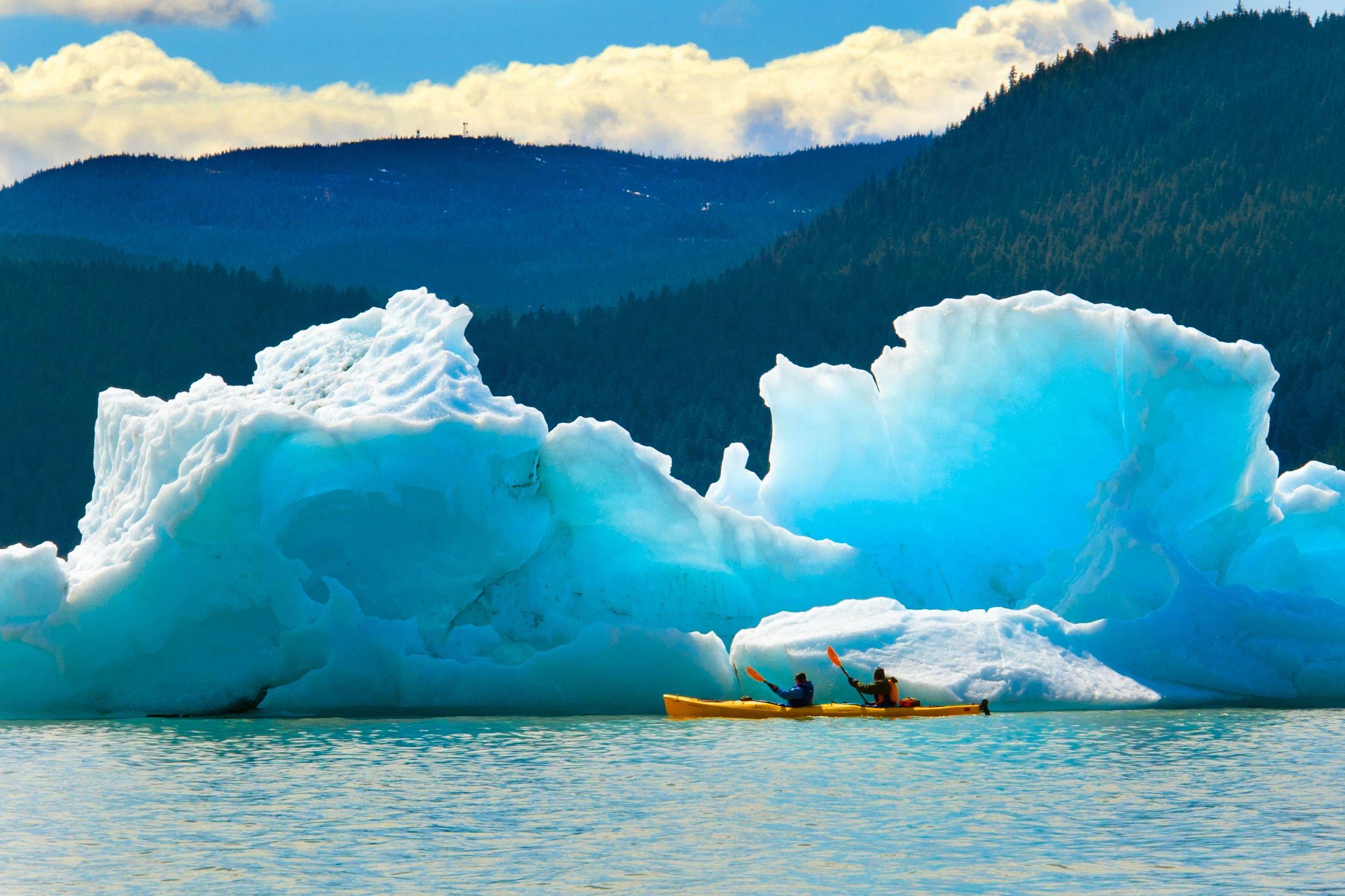 Two adventurers navigate a tandem kayak past a towering neon-blue iceberg near the Mendenhall Glacier in Juneau, Alaska.