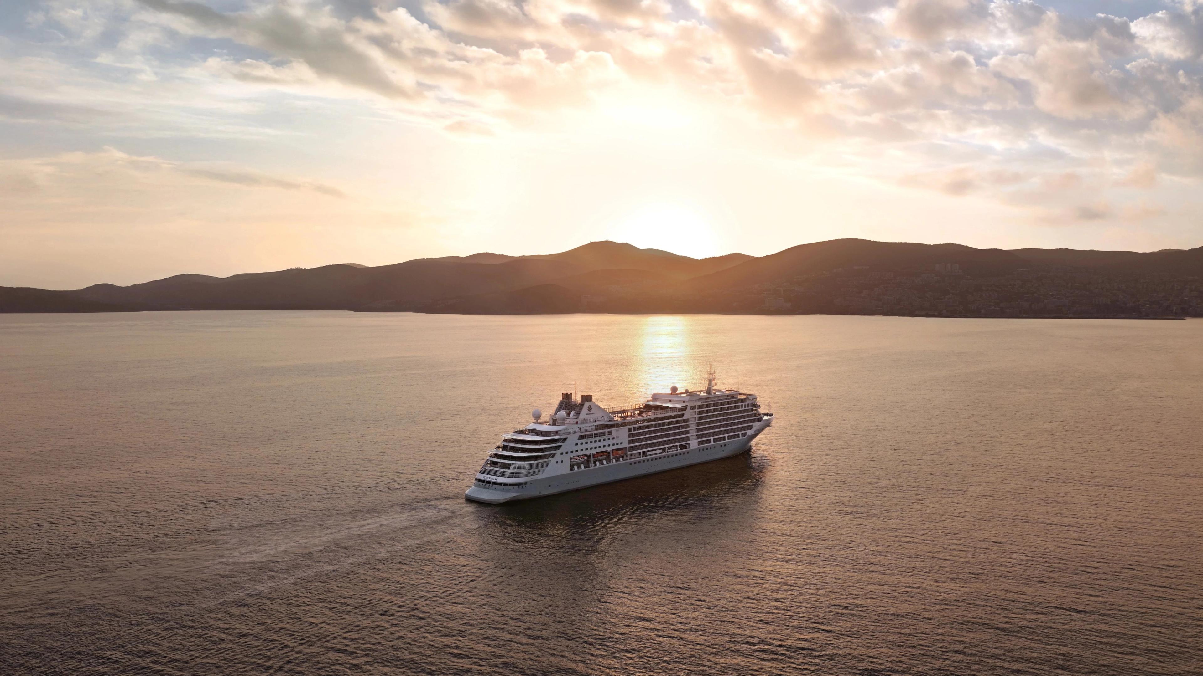 Luxury cruise ship navigating golden sunset harbor waters framing distant Kusadasi Turkey hillsides.