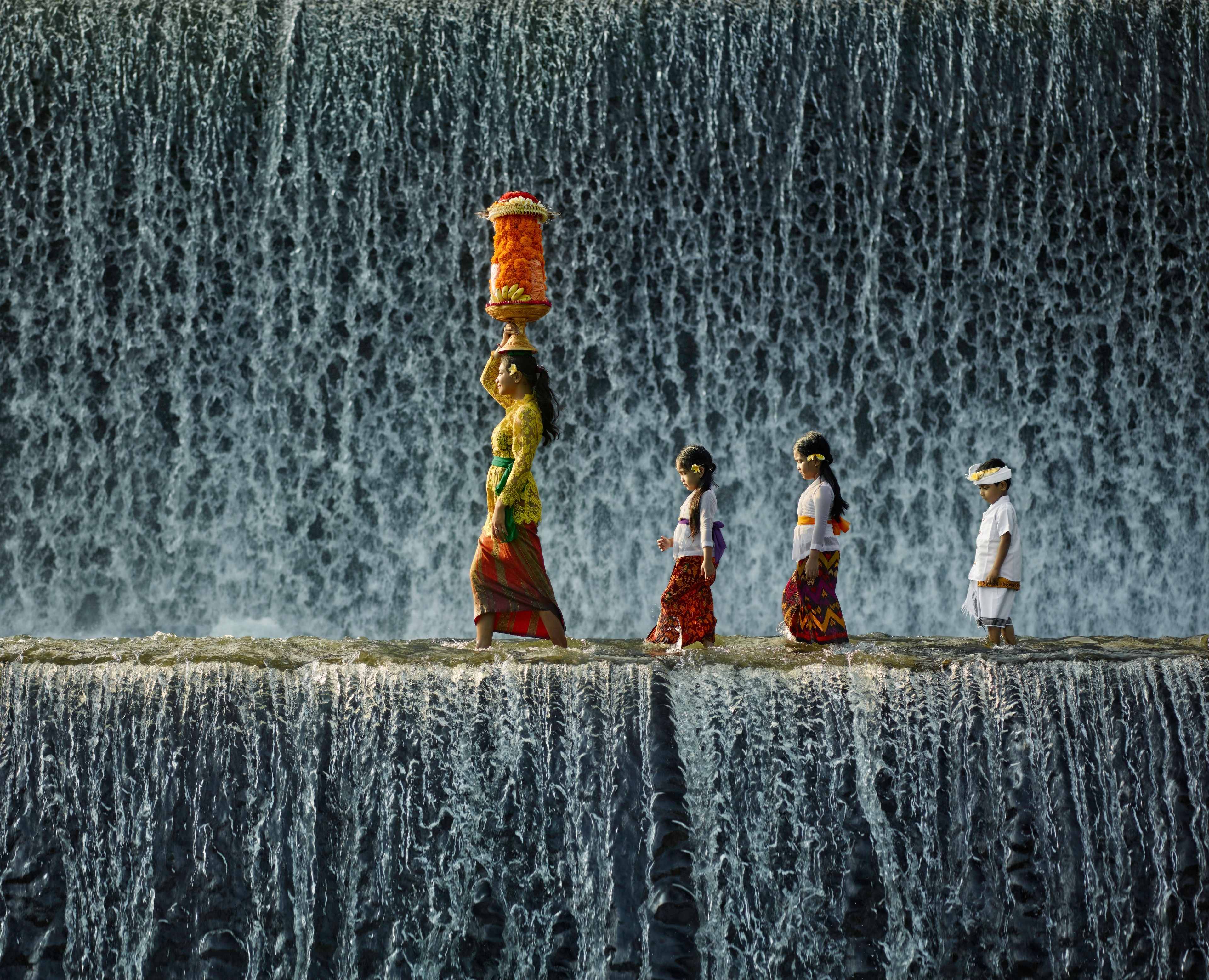 In Bali, a woman carries temple offerings, and children tag along across a waterfall in Bali/Getty images