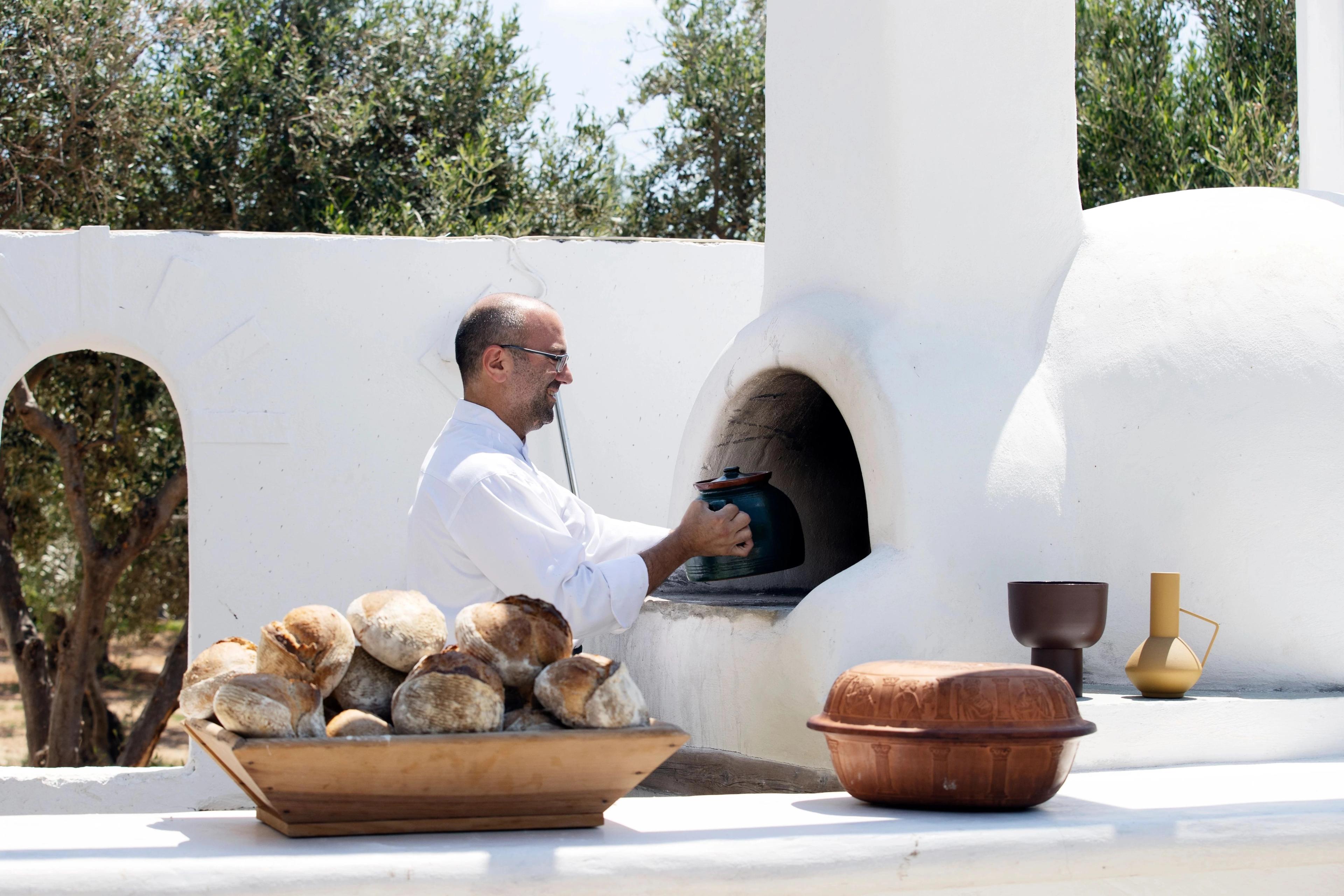 Traditional Greek sourdough bread loaves fill a rustic wooden container beside a white outdoor oven