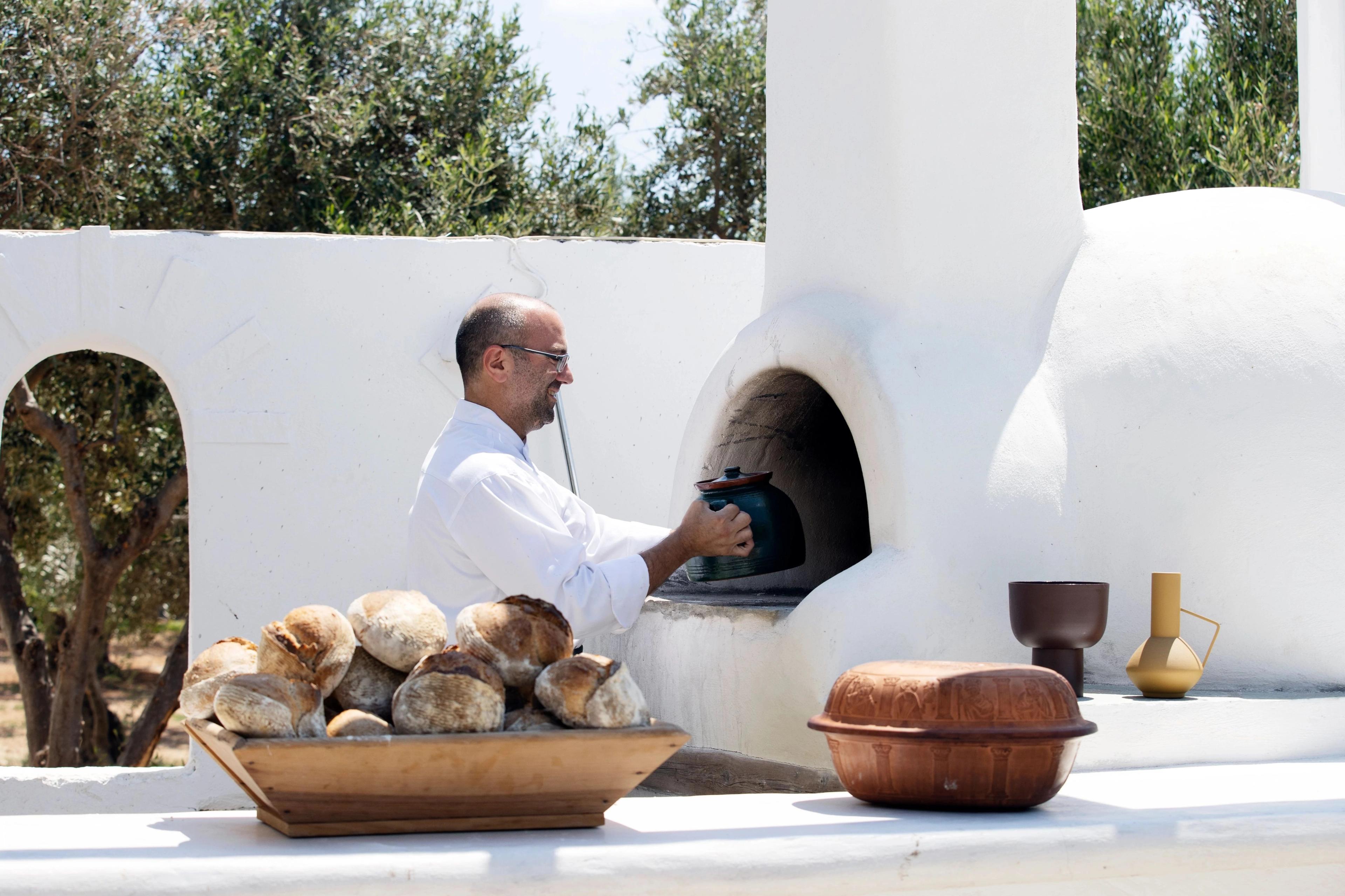 Traditional Greek sourdough bread loaves fill a rustic wooden container beside a white outdoor oven