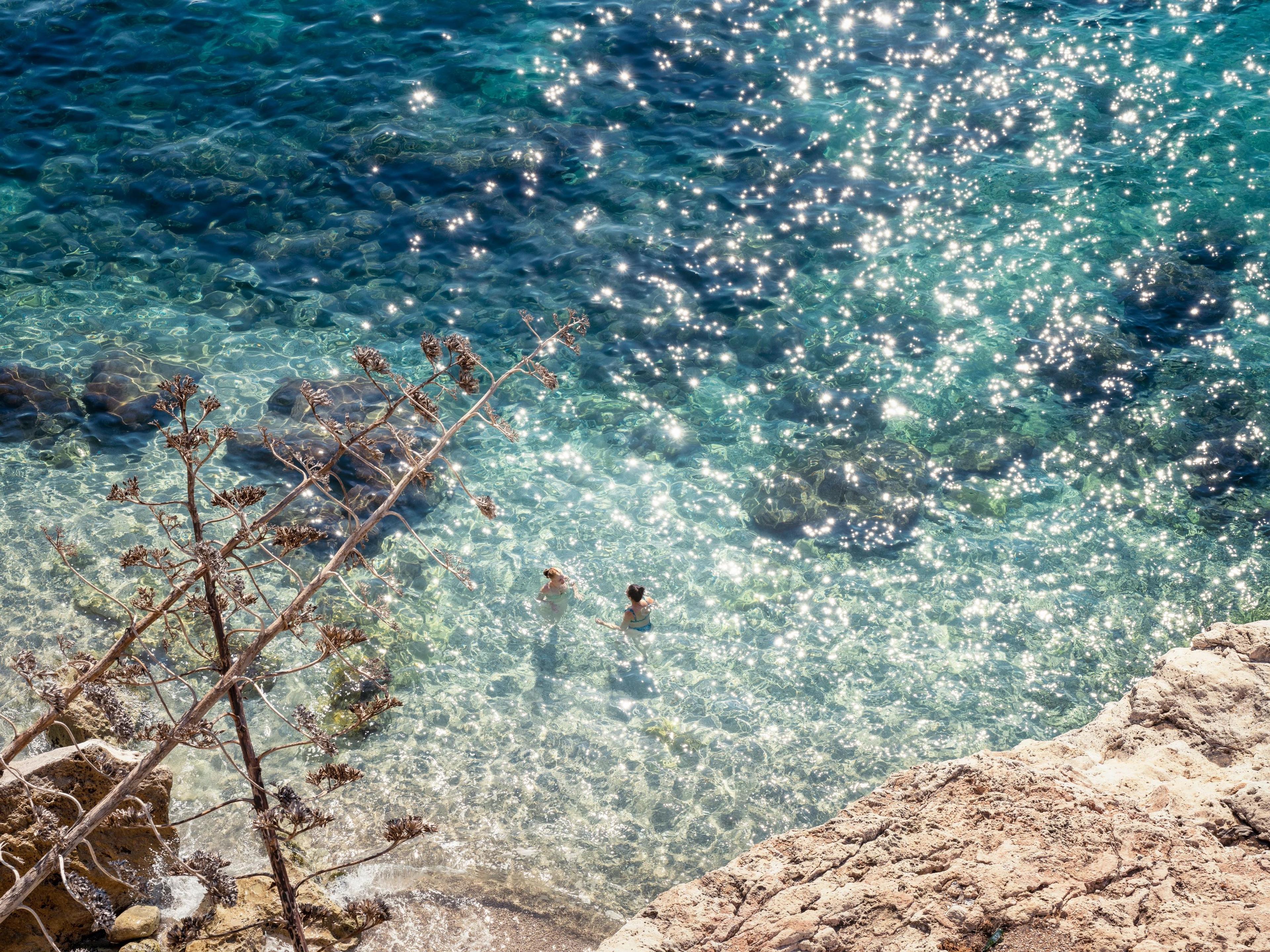 Dubrovnik Banje Beach overview showcasing sunbathers swimming beside glittering translucent turquoise Mediterranean sea waters. 