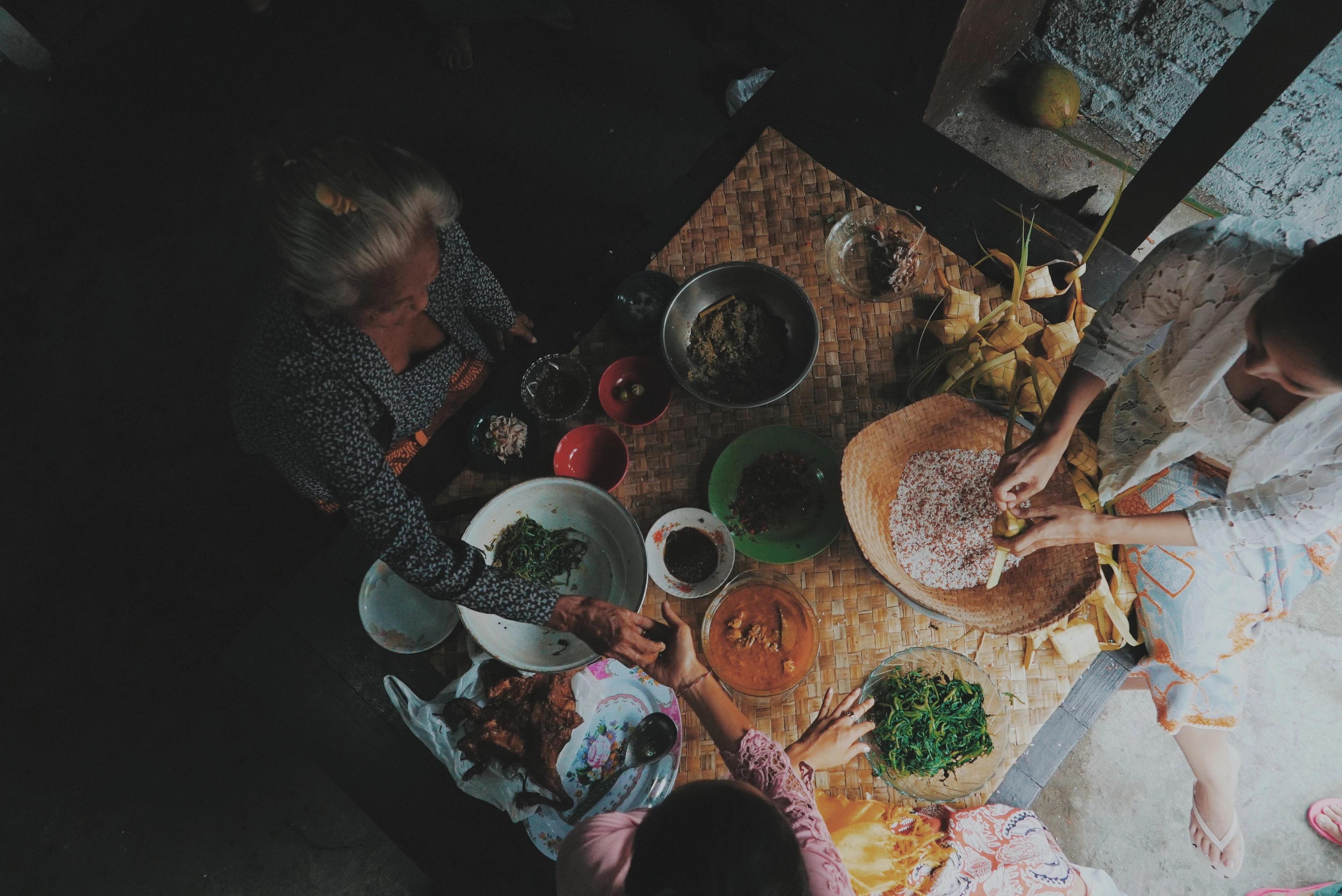 Balinese families gathering indoors to share traditional ceremonial dishes spread upon a woven bamboo mat.