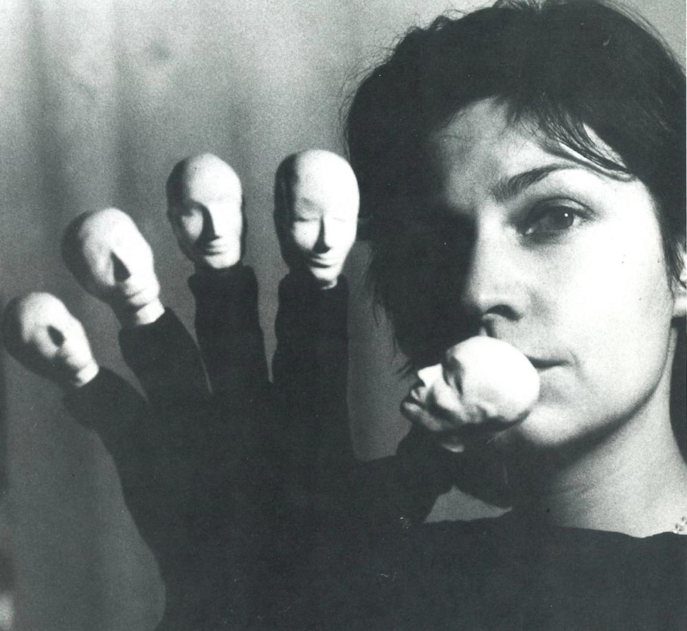 Portrait of Angelika Oehms. She holds her hand up in front of her face, with head-puppets on her fingers.