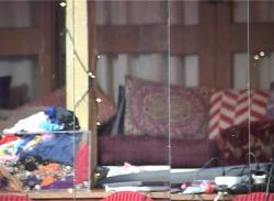 Ein Film Still zeigt die Reflektion von verschieden gemusterten Kissen, die im Hintergrund vor einer Hausfassade aufgetürmt sind. Im Vordergrund lässt sich eine Tischplatte erahnen, auf der viele Kabel, Papiere, eine durchsichtige Kiste mit Textilien und weiterer Kram liegt.