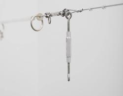 im Vordergrund ist ein quer-gespannter Drahtseil zu sehen, welches durch an ihr befestigte verschiedene Arten von Ringen, Muttern, Haken, sogenannte Hardware, aufgespannt wird. Hintergrund ist verschwommen.