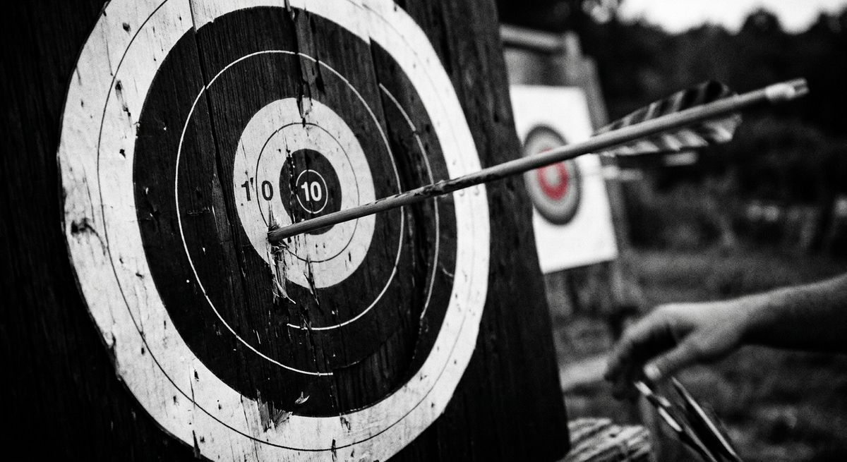 Black and white photograph of an arrow embedded off-center on a target, near the edge.