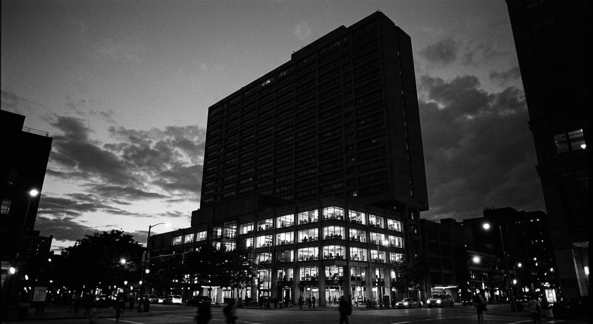 Black and white photograph of a large building at dusk with only the lower floors lit and the upper floors dark.