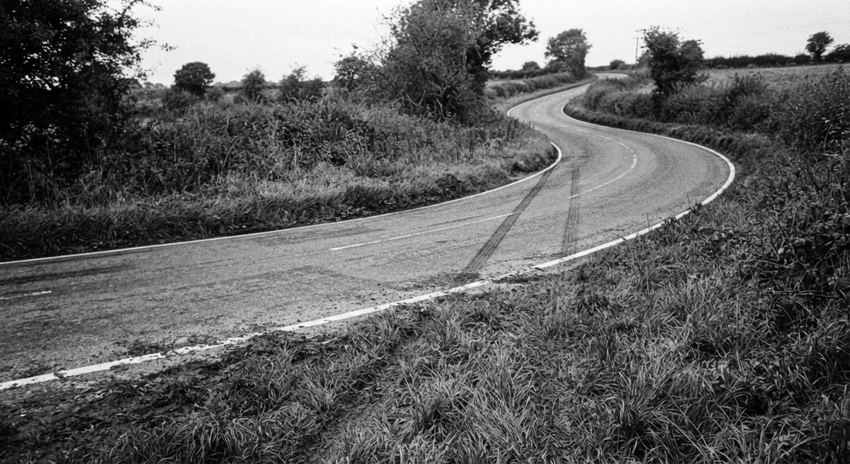 Black and white photograph of a road curving sharply while a set of straight tire tracks continues off the edge.