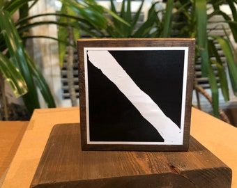 A framed print of a squared Diagolon flag. It sits on a stained wooden surface and large plants are behind it.
