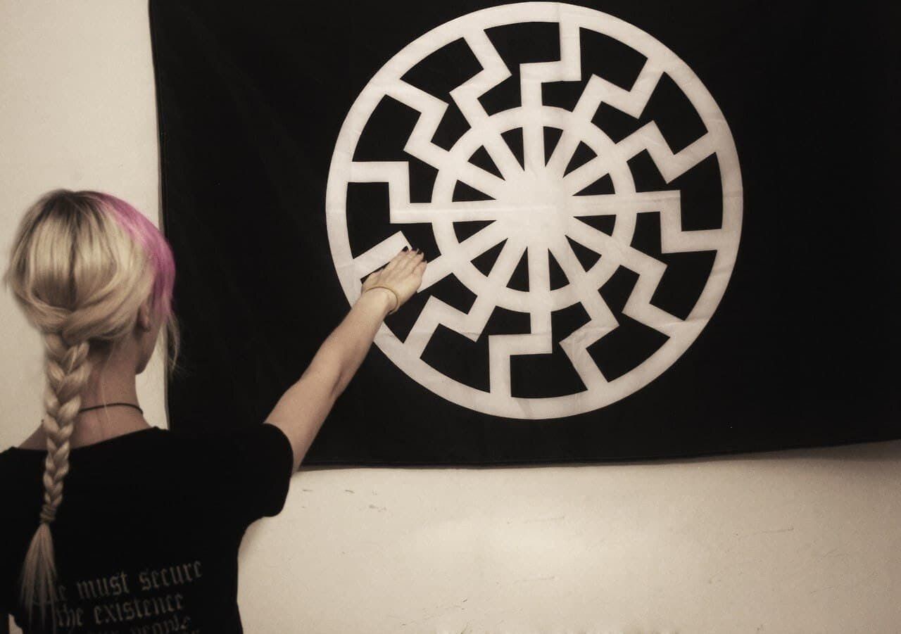 Photograph. A women with blond and pink hair in a braid gives a Nazi salute to a black flag with a white 'Black Sun' on it. On the back of her shirt is the Fourteen Words in a blackletter typeface.