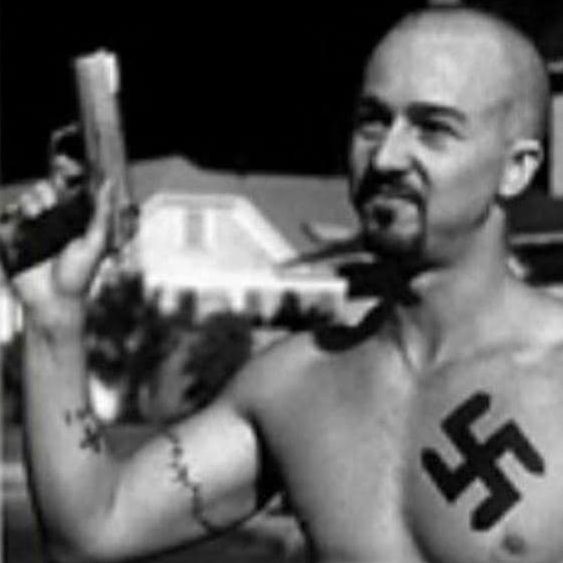 A still image from American History X showing Derek Vinyard (portrayed by Edward Norton) with a swastika tattoo and a pistol.