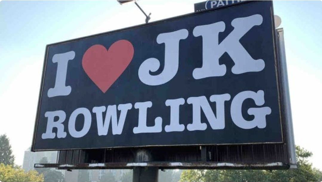 Photographie d'un panneau publicitaire. Le texte à empattement en bloc blanc indique "I (coeur) JK ROWLING" sur fond noir.