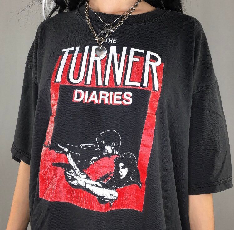 Photographie d'une femme portant un t-shirt avec la pochette de Turn Diaries imprimée dessus. L'art représente un homme et une femme tirant des armes à feu sur des cibles hors de vue.